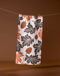 The Geometry Woodland Waltz bar towel, made from recycled materials, features an autumn-inspired pattern of orange and black leaves with pinecones and hangs on a clothesline against a brown background.