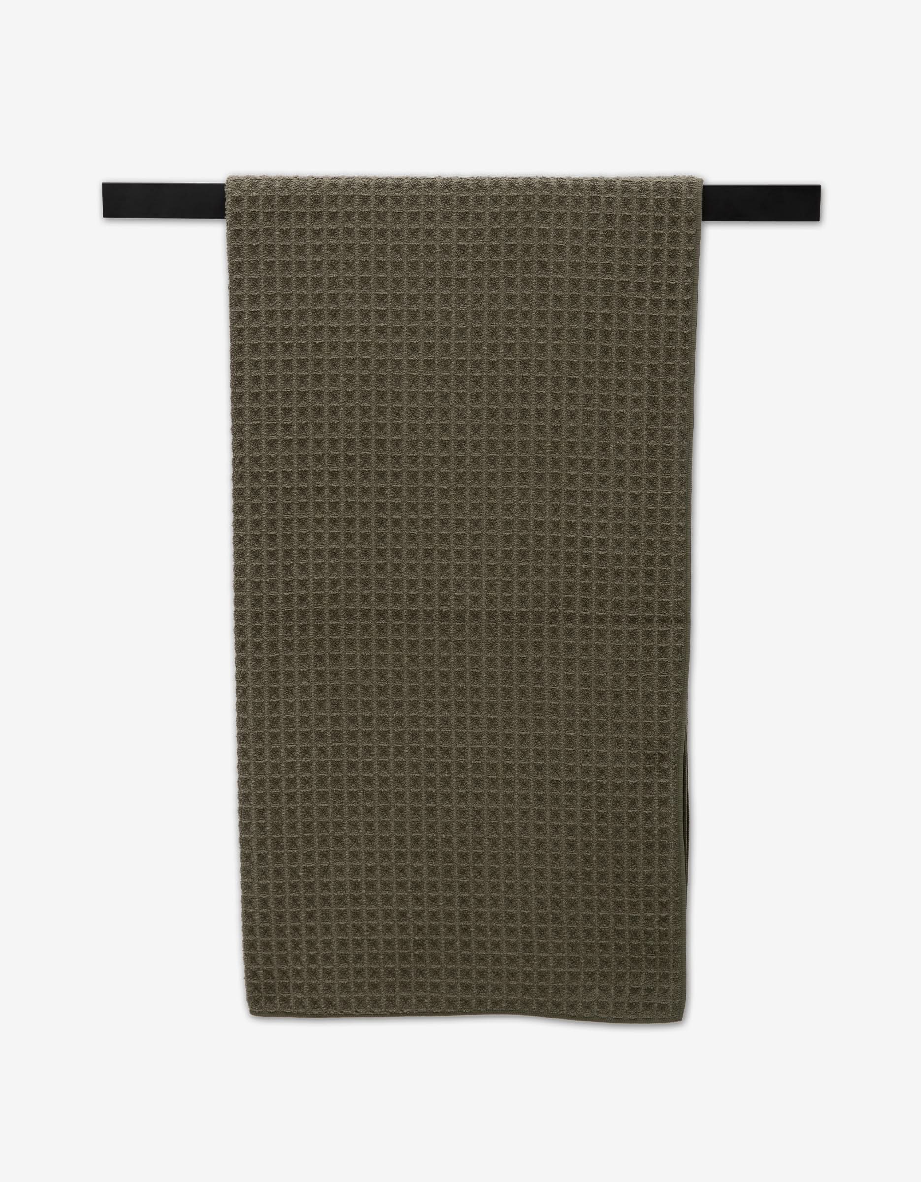 The Forest waffle bath towel by GEOMETRY in dark olive green hangs on a black towel bar against a white background, adding stylish luxury to your space.