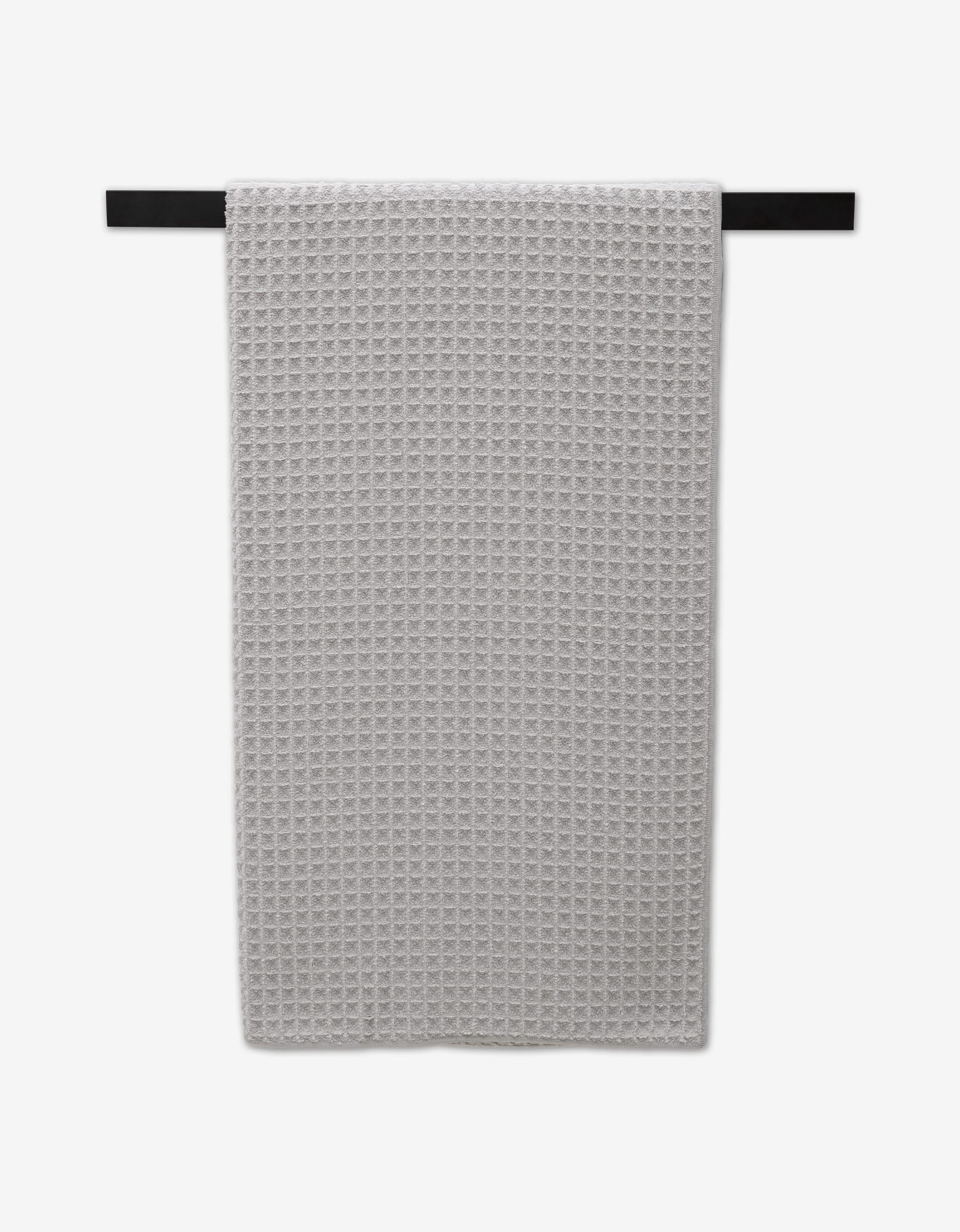 A GEOMETRY Stone ultra-absorbent Waffle Bath Towel in light gray, featuring a textured waffle pattern, hangs neatly on a black horizontal bar against a plain white background.