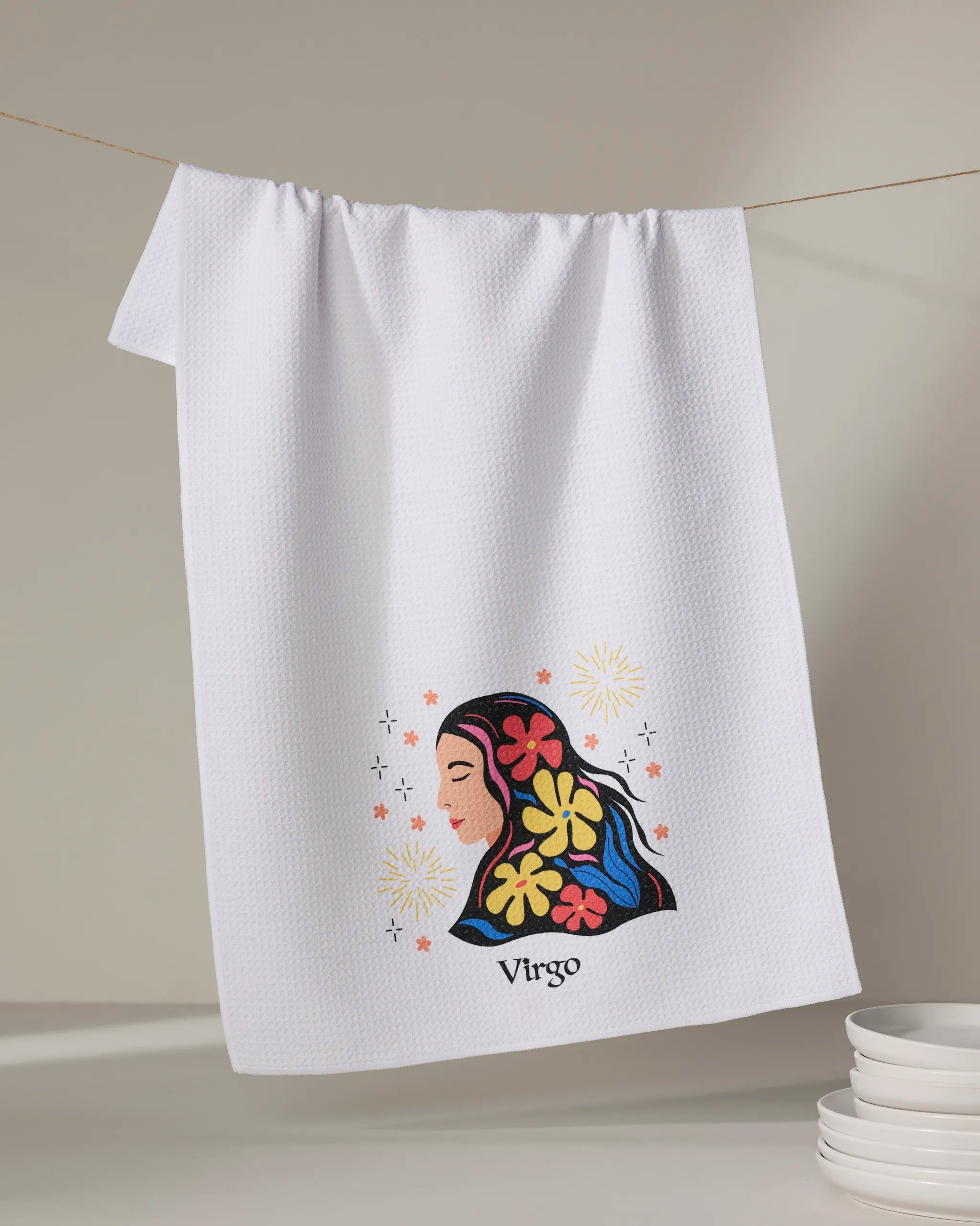 A white textured kitchen towel from Heather Zhou, called "Clever Virgo," hangs on a line. It showcases a colorful embroidered woman's face with flowers and stars, "Virgo" below, and stacked white dishes in the lower right corner.