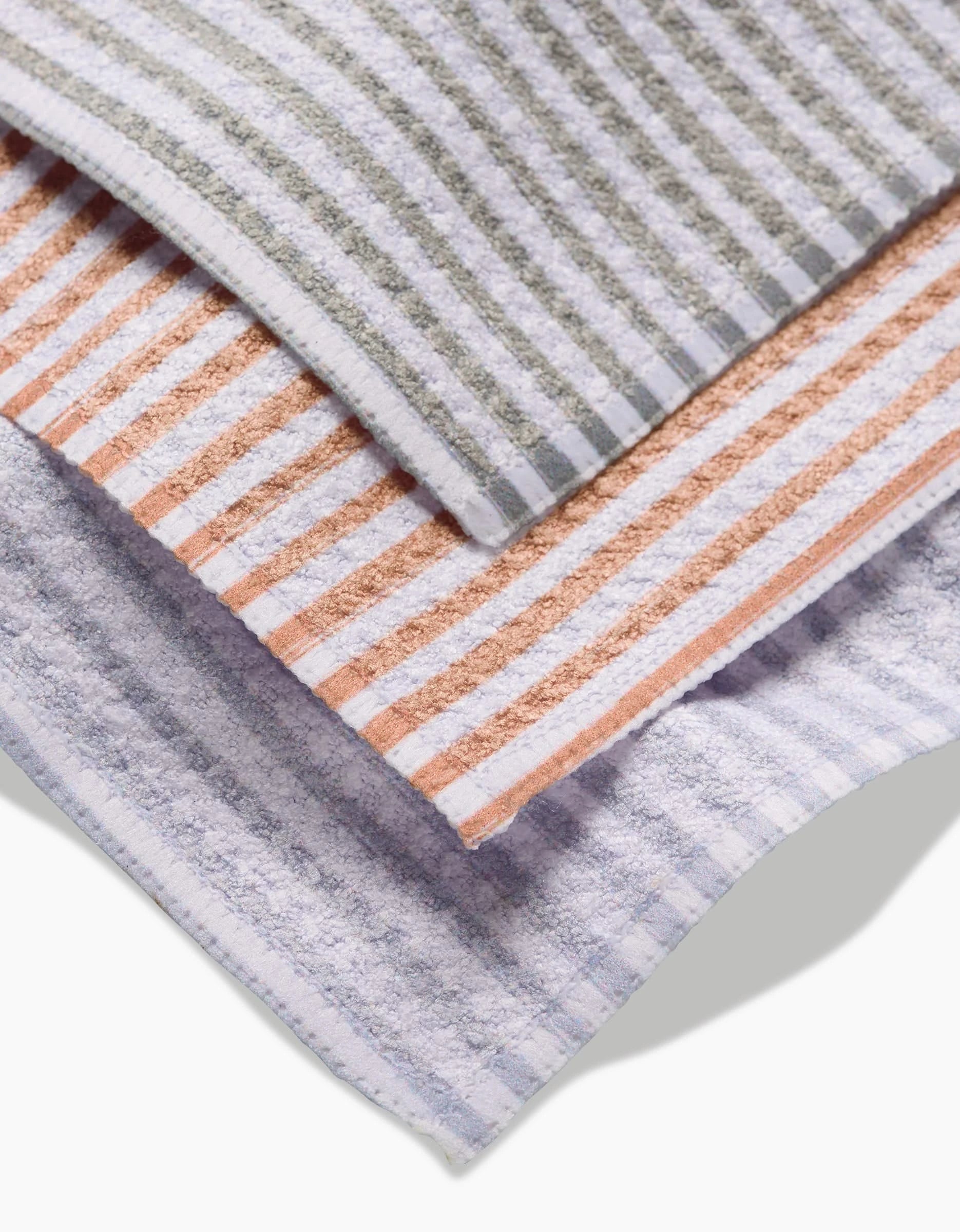 Three À La Carte towels by Kailee Wright, made from recycled materials, are stacked: the top has gray and white stripes, the middle is orange and white, and the bottom features light purple and white stripes.