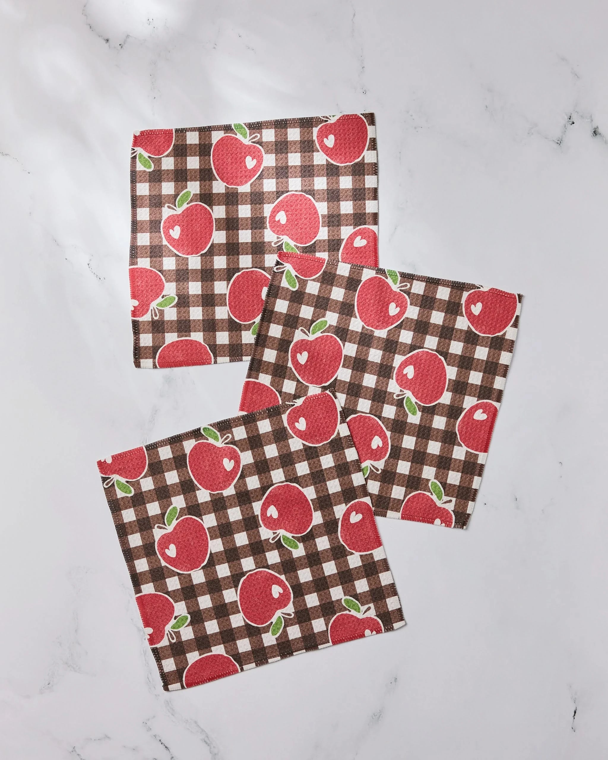Three brown and white checkered napkins from Geometry’s Apple Picking Afternoon set, featuring red apples with green leaves, are arranged on a white marble surface. Made from recycled materials, they create a cozy kitchen display.