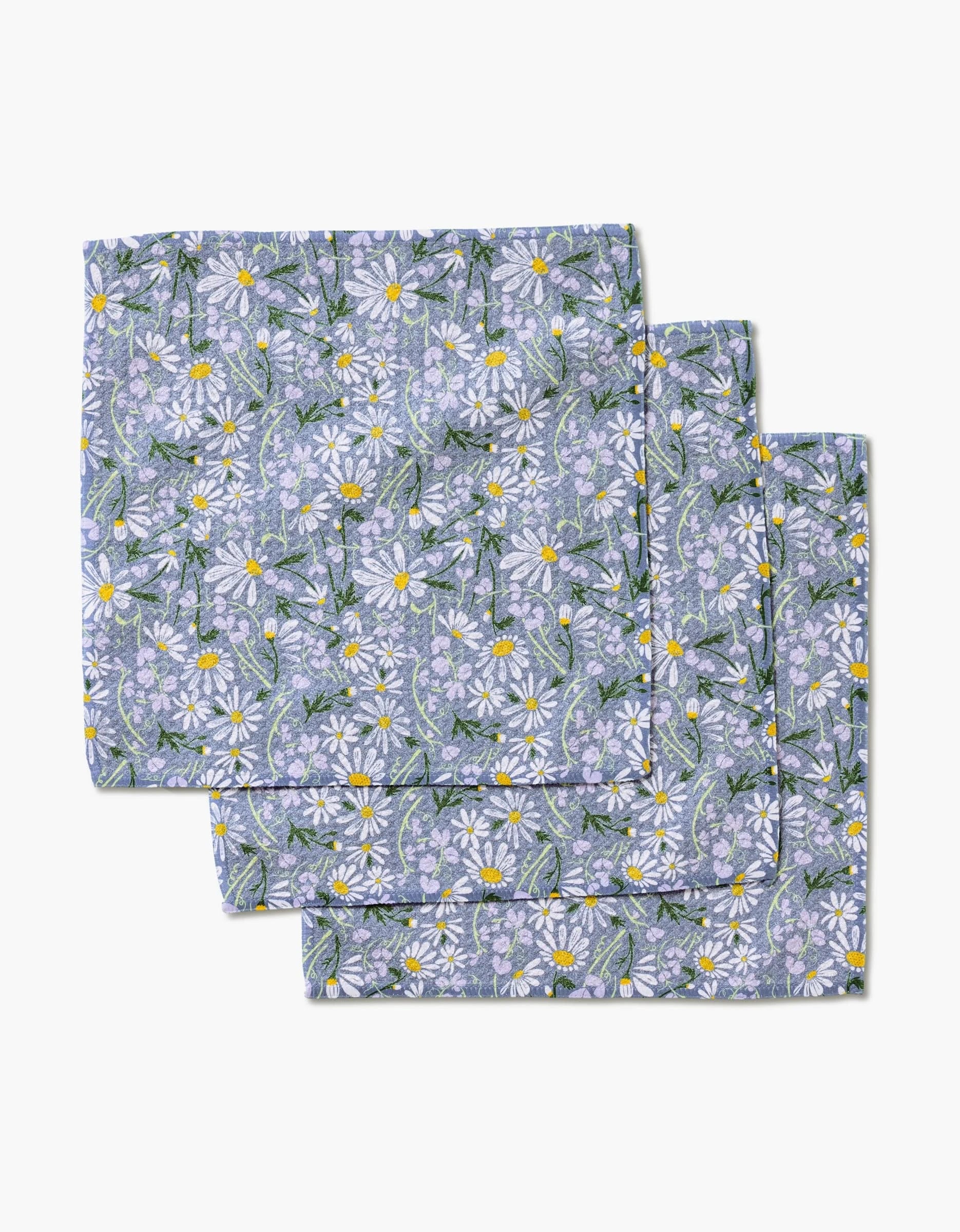 Three April napkins by Elisabeth Wing, crafted from eco-friendly Geoweave fabric, display white daisies with yellow centers and green leaves on a purple background, shown slightly overlapping on a white surface.