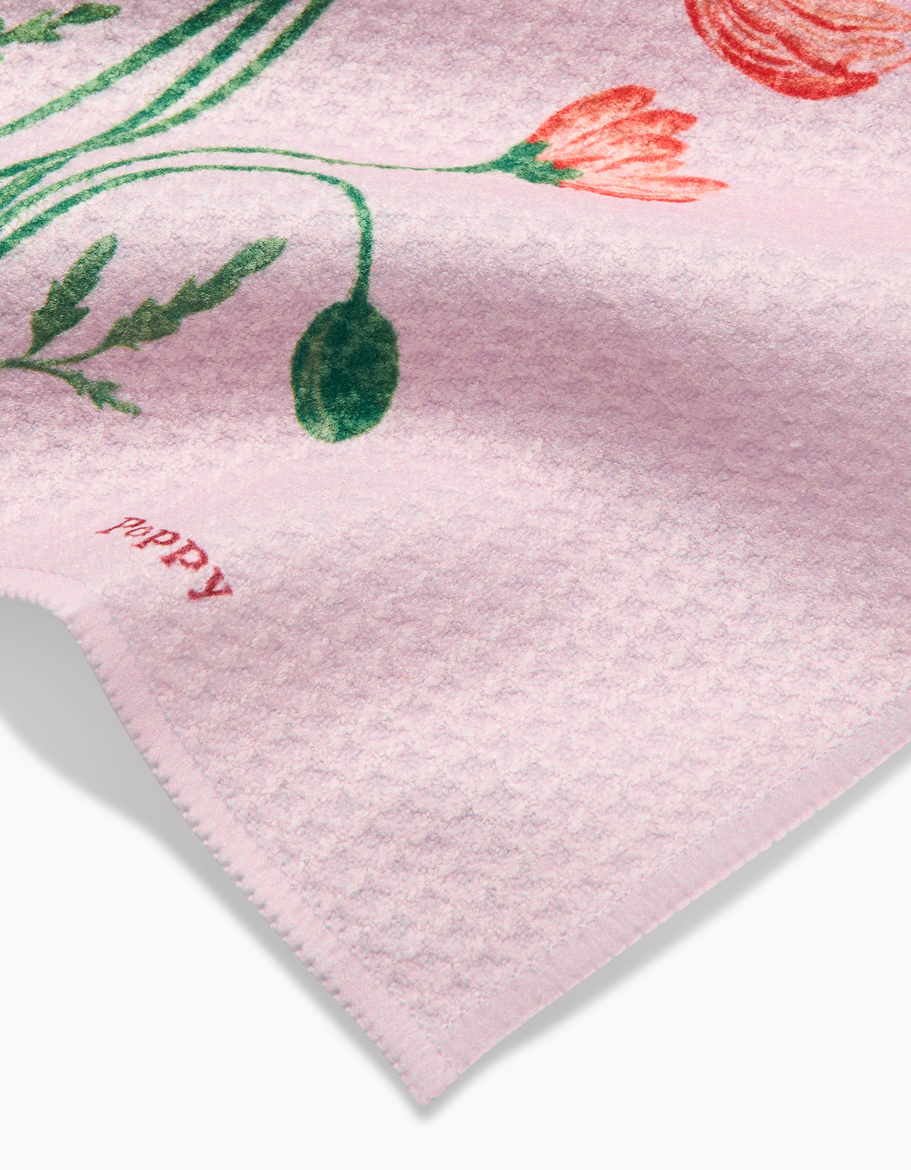 A close-up of the August Poppy dishcloth by Linda Fox, made from soft pink Geoweave fabric, shows green leaves and red flowers with poppy in small red letters near the edge.