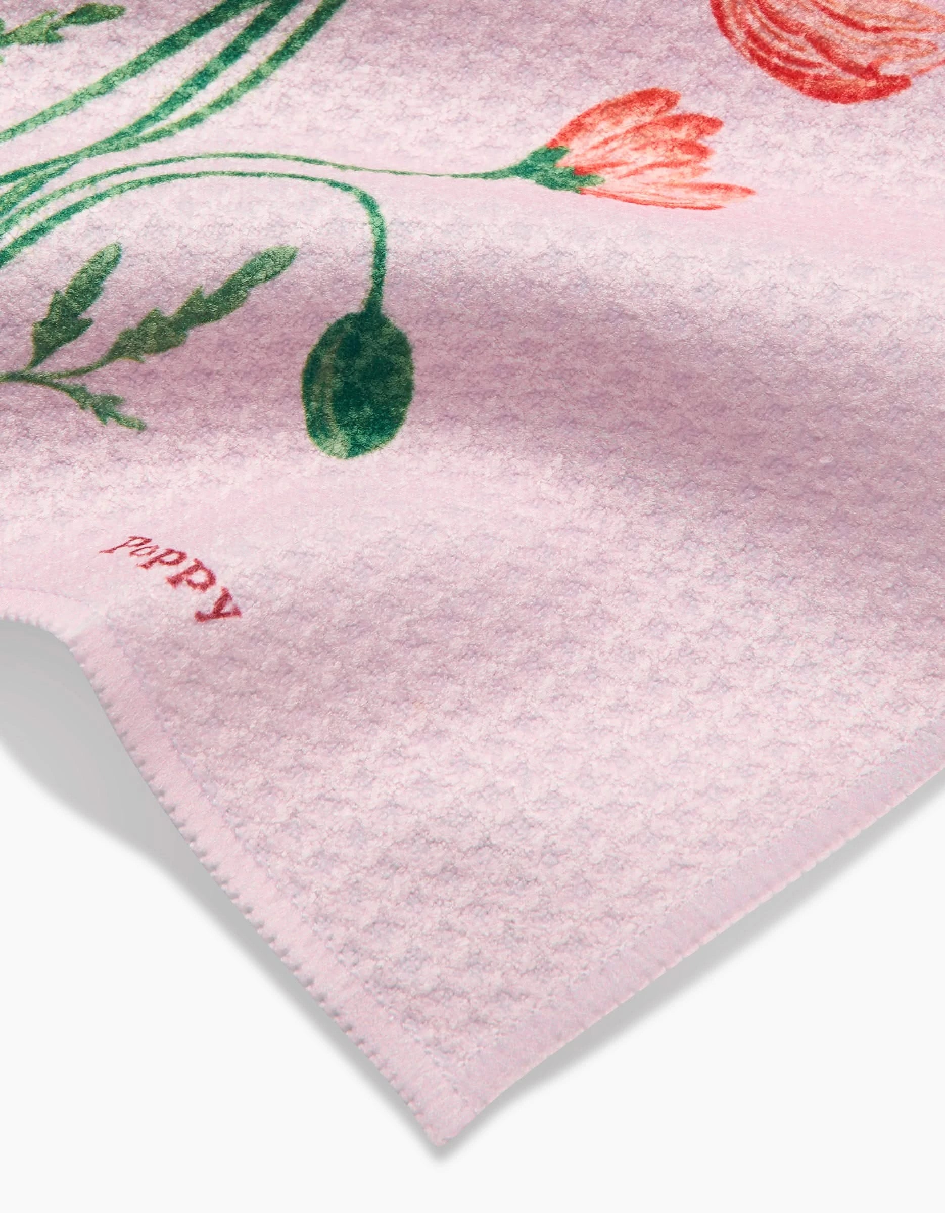 A close-up of the August Poppy dishcloth by Linda Fox, made from soft pink Geoweave fabric, shows green leaves and red flowers with poppy in small red letters near the edge.
