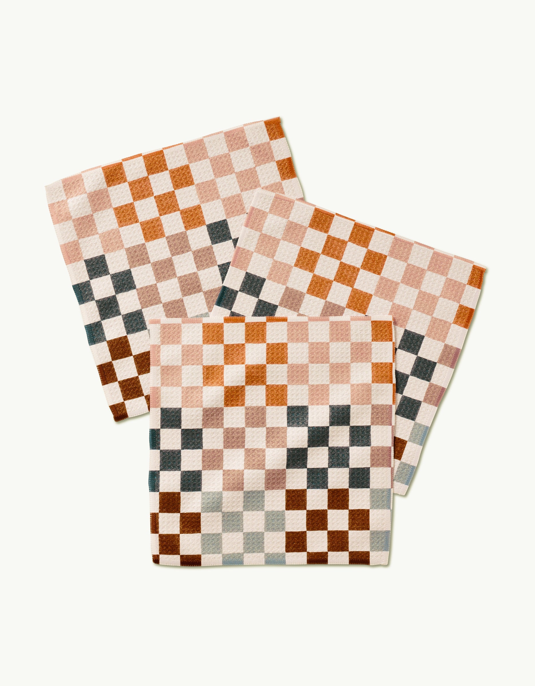 Three Autumn Checkers cloth napkins by GEOMETRY, made from recycled materials and featuring a cream, brown, pink, rust, and dark gray checkered pattern, are overlapped on a light background.