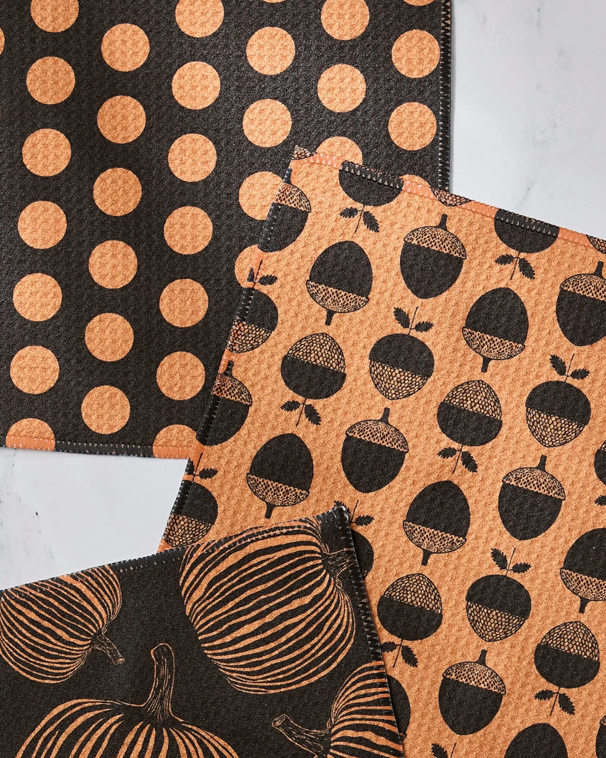 Three Geometry Autumn Noir cloth napkins—one with orange polka dots on black, one with black acorns on orange, and one with black pumpkins on orange—are shown on a light surface. Each is made from recycled materials.