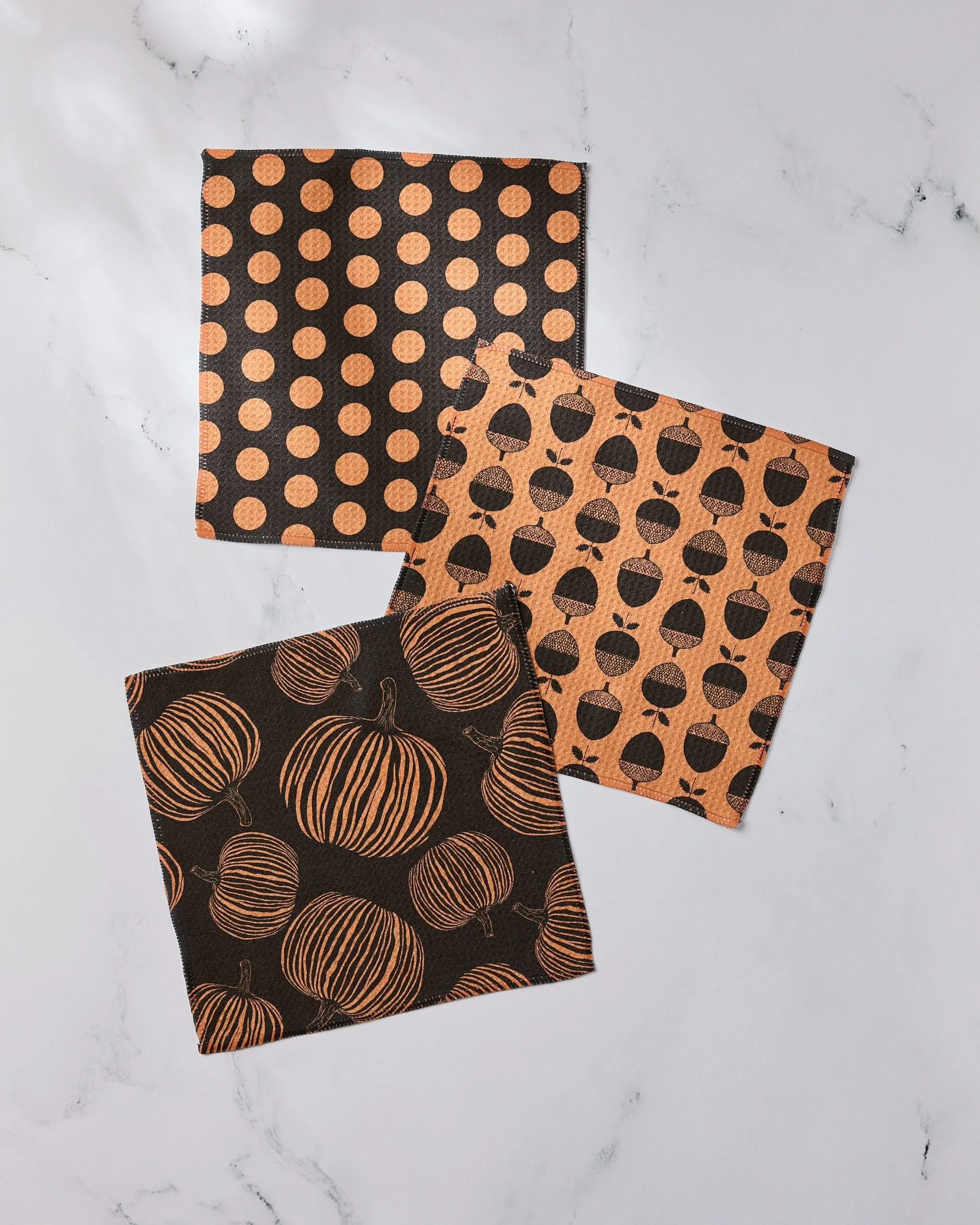 Autumn Noir by Geometry: Three square napkins made from recycled materials feature Halloween patterns—orange polka dots on black, black pumpkins on orange, and orange pumpkins on black—displayed on white marble.