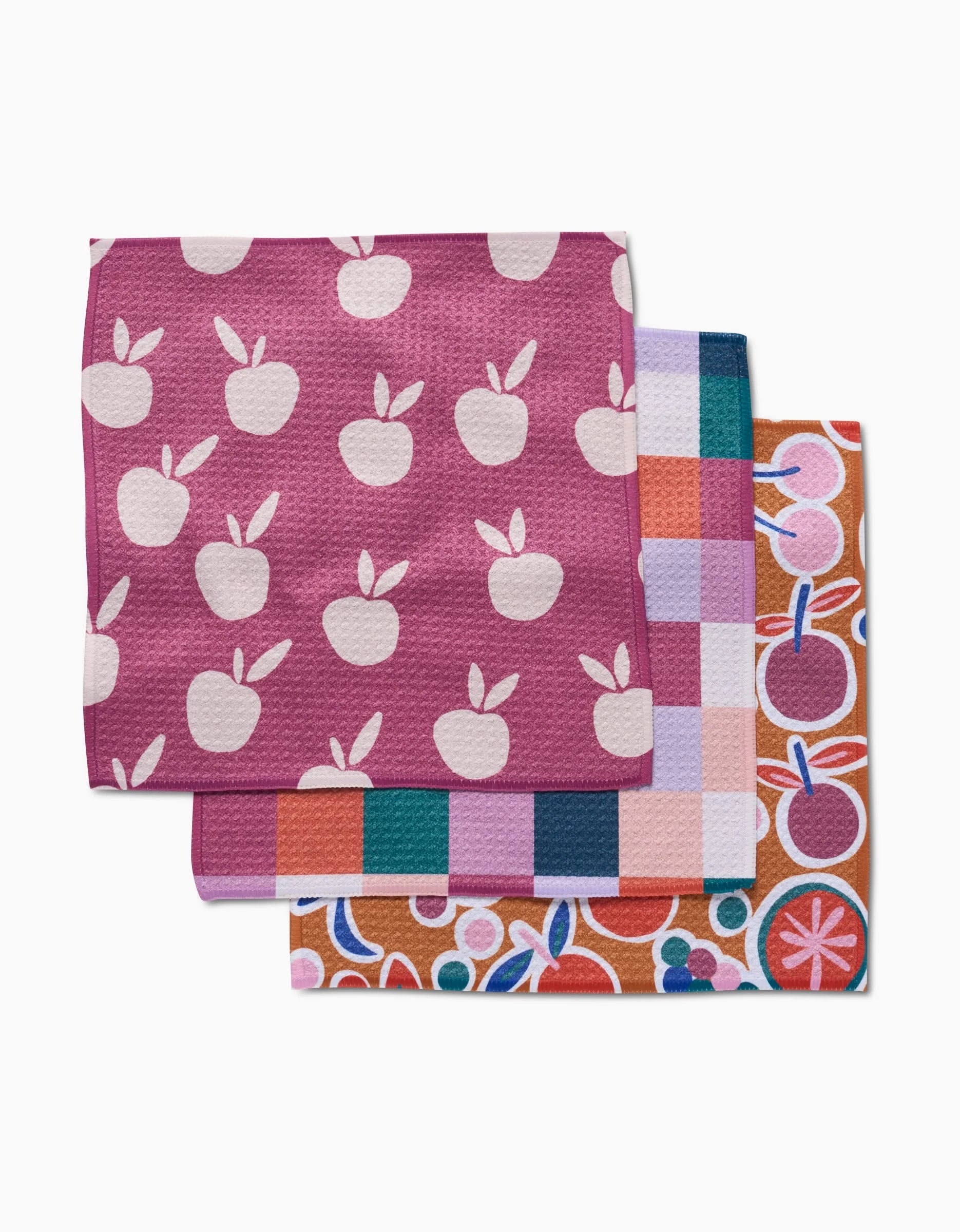 The Claire Iglesias Autumn Party set includes three eco-friendly kitchen towels with vibrant patterns: pink with white apples, multicolored checks, and orange with fruit and floral designs.