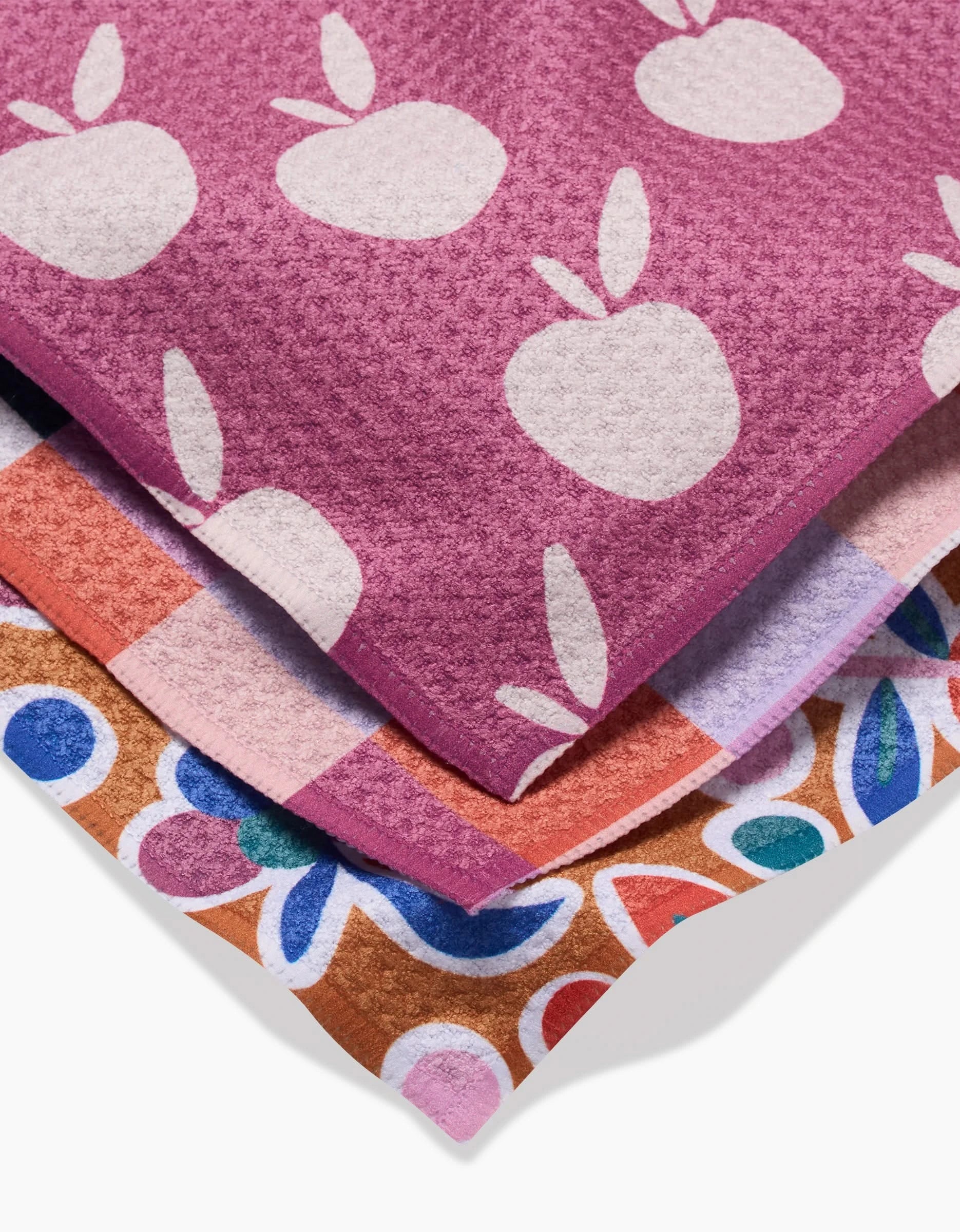 Three eco-friendly kitchen towels from Claire Iglesias’s Autumn Party set are stacked: a pink towel with white apples, one in a pink, purple, and white checkered design, and another with a bold multicolor floral pattern.