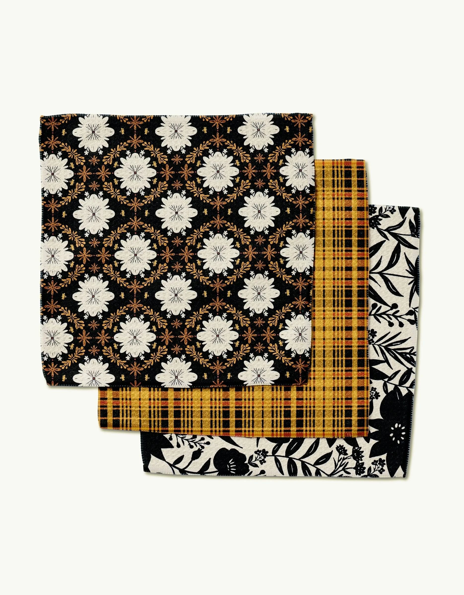 Three folded Danelys Sidron Autumn Plaid napkins are stacked: the top has a black and white floral motif with orange accents, the middle features a yellow and black plaid, and the bottom shows a black and white botanical design.