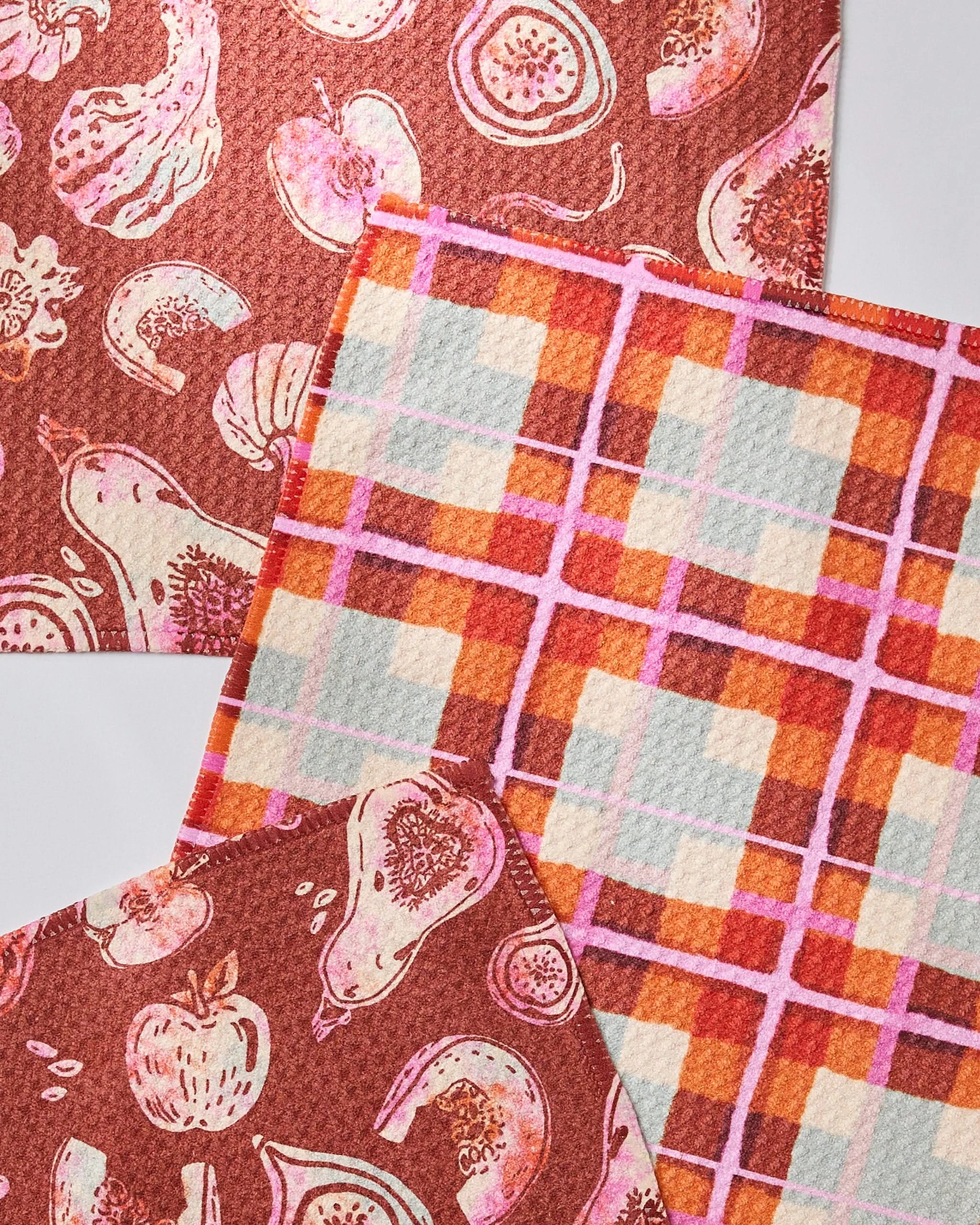 The Geometry Autumnal Love set includes three eco-friendly Swedish dishcloths: two with reddish-brown backgrounds and pink produce designs, plus one plaid in orange, red, pink, and beige—each made from recycled materials.