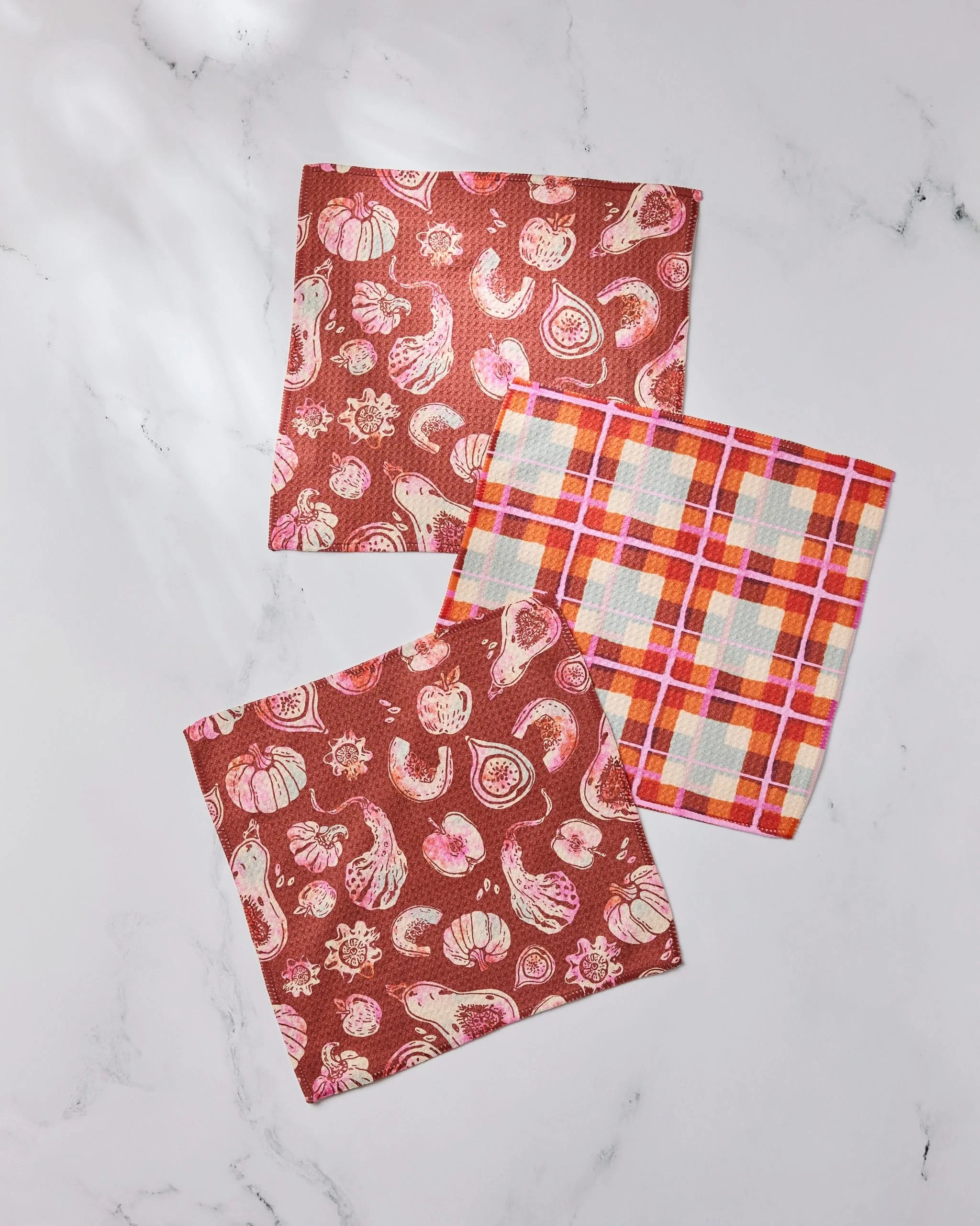 Three Geometry Autumnal Love dishcloths on a white marble surface: two with red backgrounds and pink-white veggies, and one with a bold red, orange, and white plaid pattern.