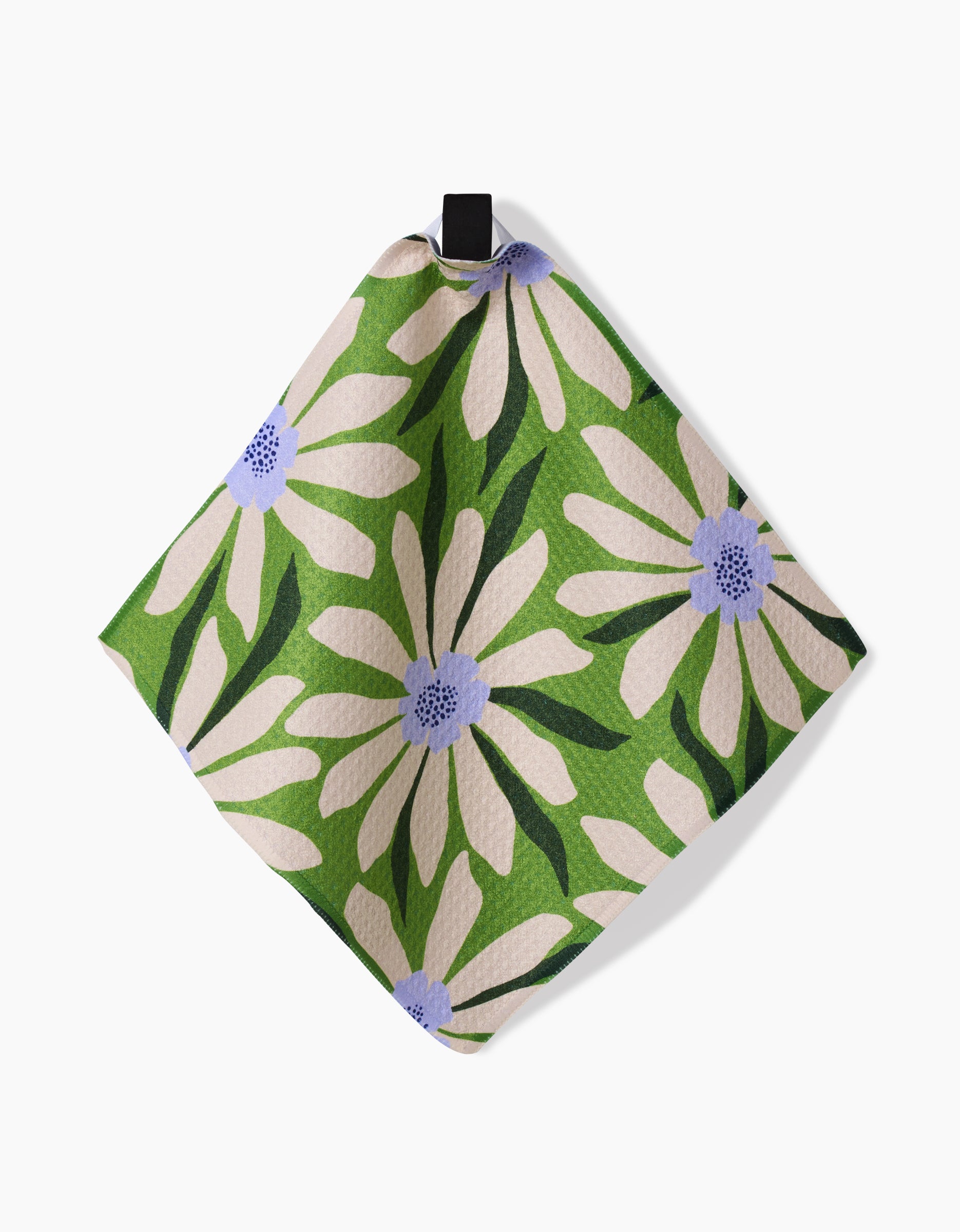 The GEOMETRY Bliss and Bloom napkin features textured Geoweave fabric, a green background with large white flowers, light purple centers, dark green leaves, and hangs by a black loop.