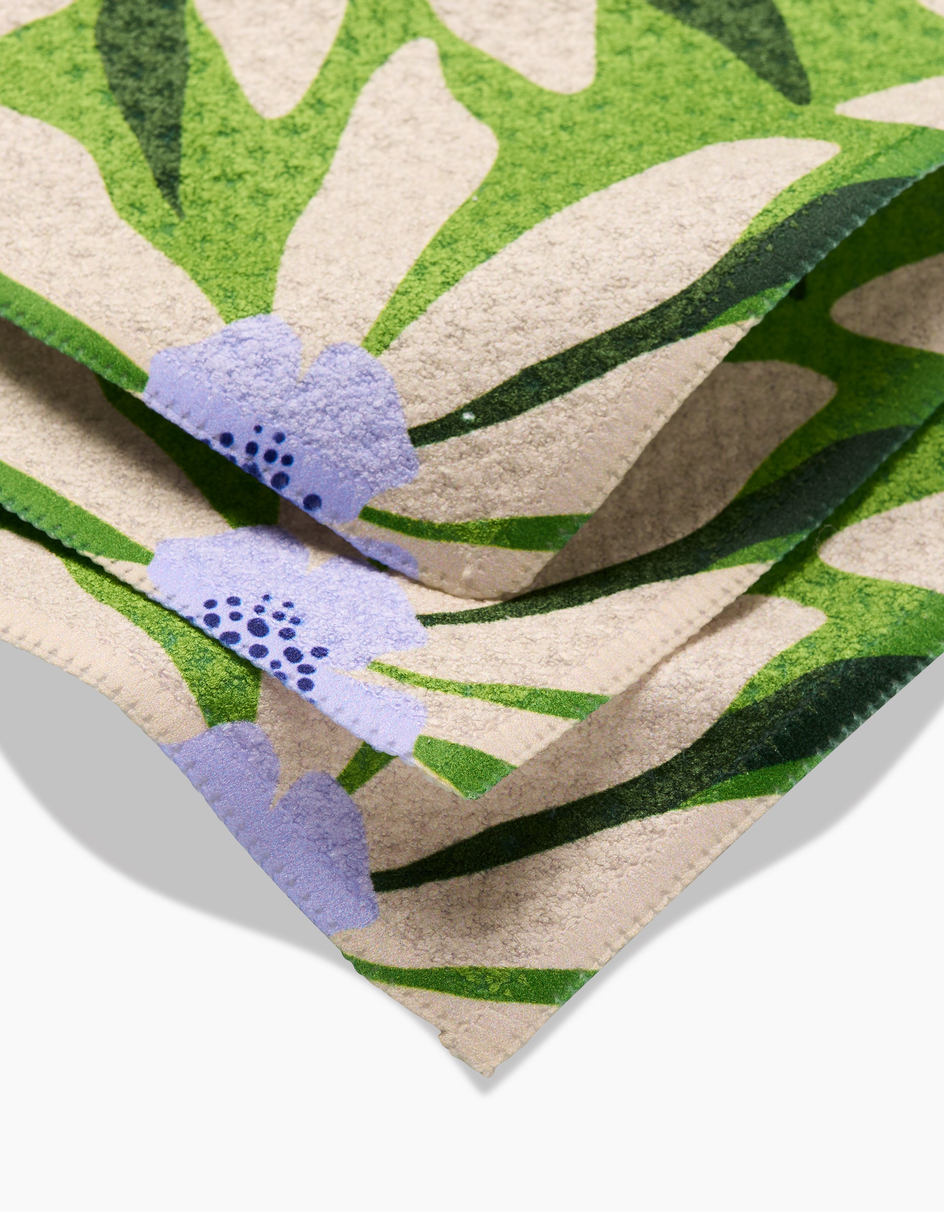 A close-up of GEOMETRYs Bliss and Bloom towels, featuring a botanical pattern of green leaves and light purple flowers on beige. Made from recycled materials, the soft-textured fabric adds an eco-friendly touch.