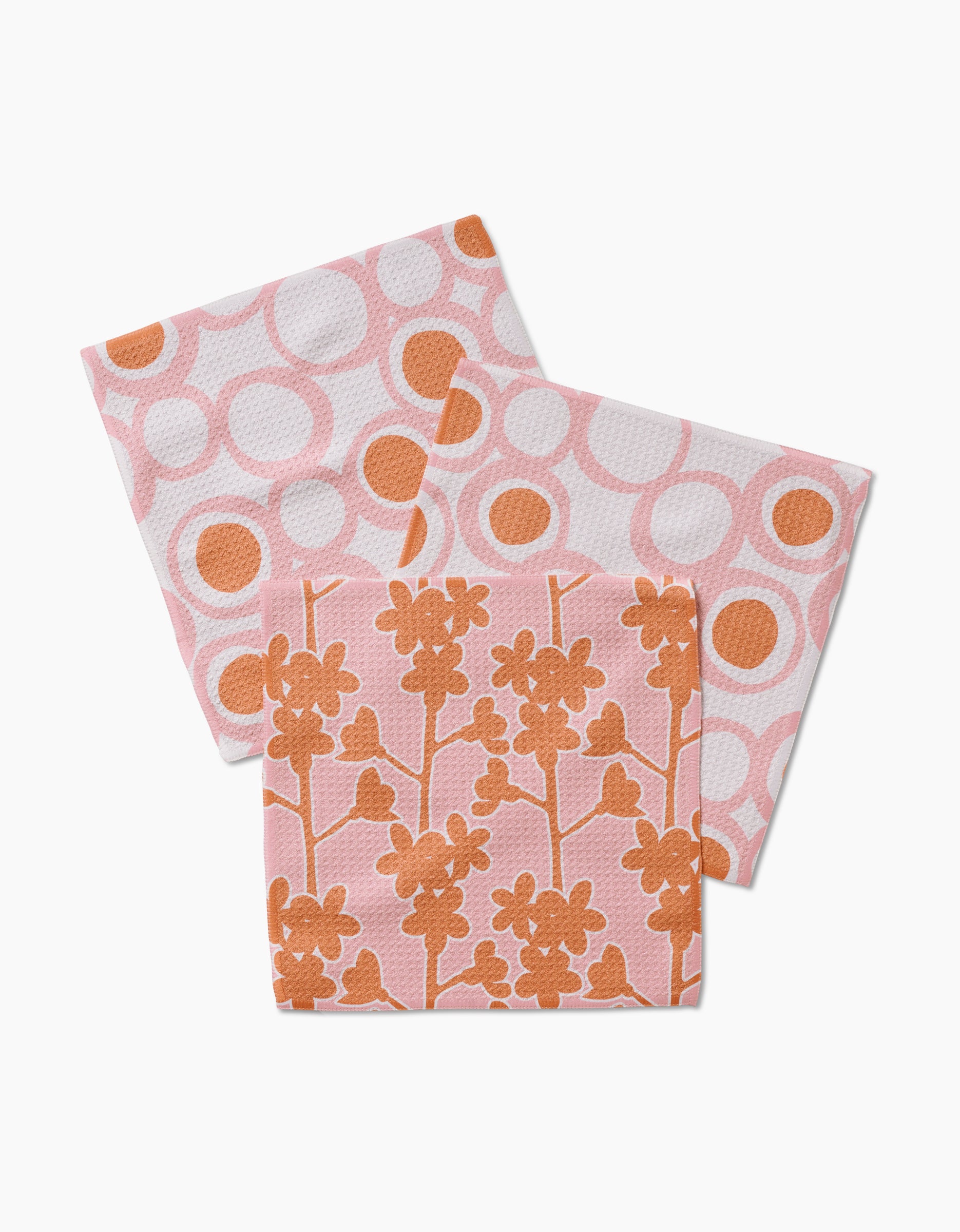 Three Bouncing Balls reusable dishcloths by Lizzie Clark are shown: two with a pink and orange circle pattern, and one with an orange floral design on a pink background, all made of Geoweave fabric on a white surface.