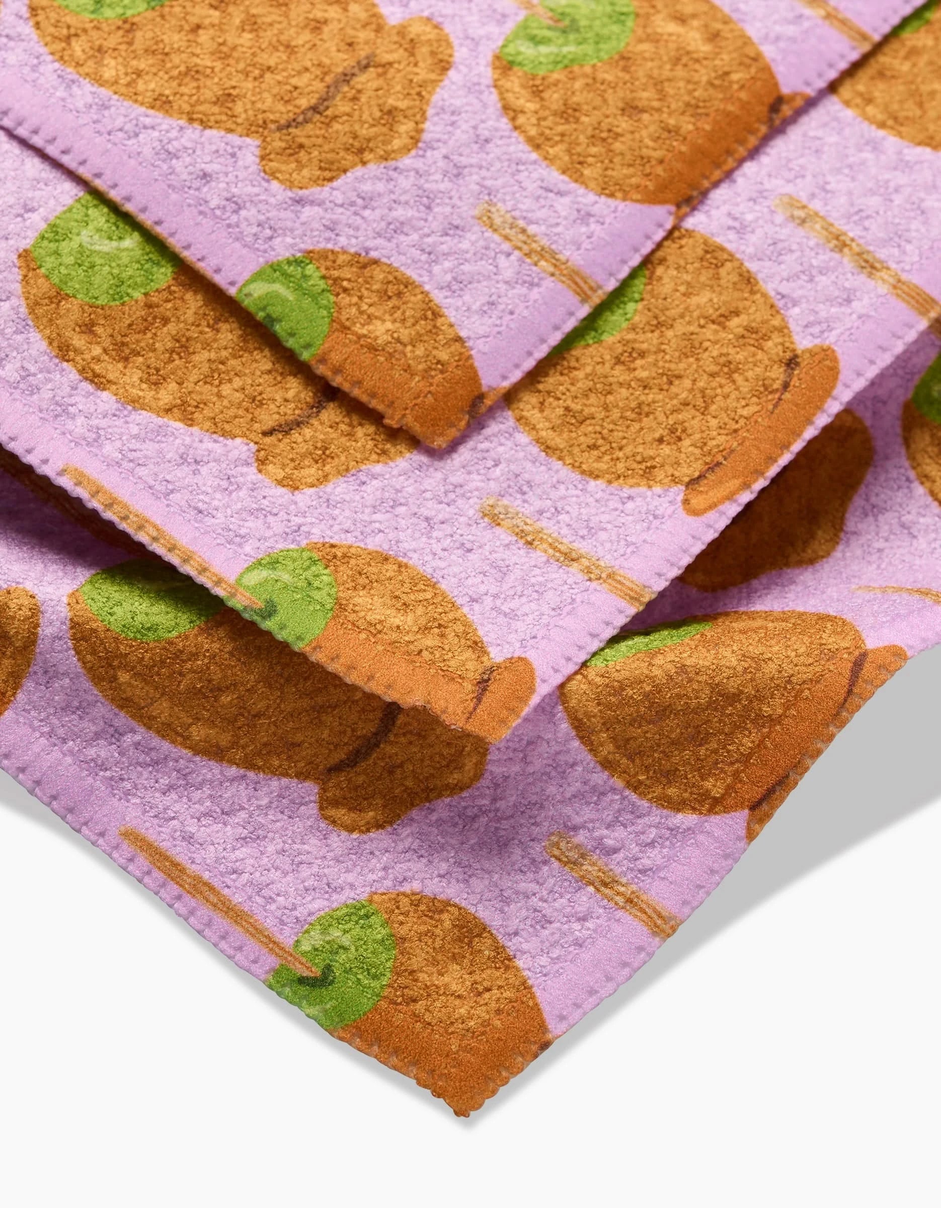 Close-up of Elisabeth Wings Caramel Apple towels, made from recycled materials with a playful brown snail shell and green head pattern on a light purple background. Towels show visible stitched edges.
