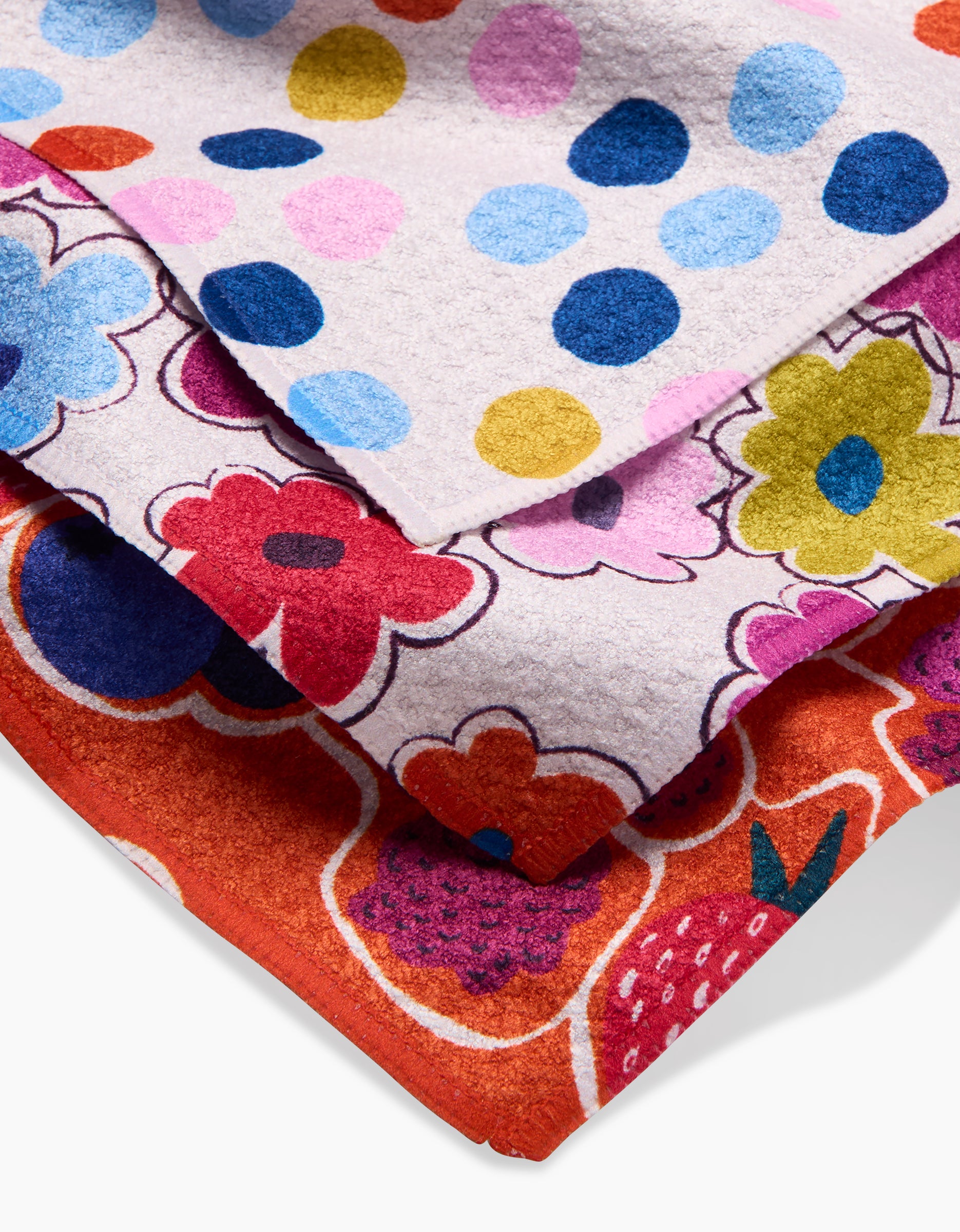 Three Carnival Fling Geoweave dishcloths by Lizzie Clark are stacked: the top has blue, pink, and yellow polka dots; the middle features multicolored flowers; the bottom displays a red background with assorted berry and fruit designs.