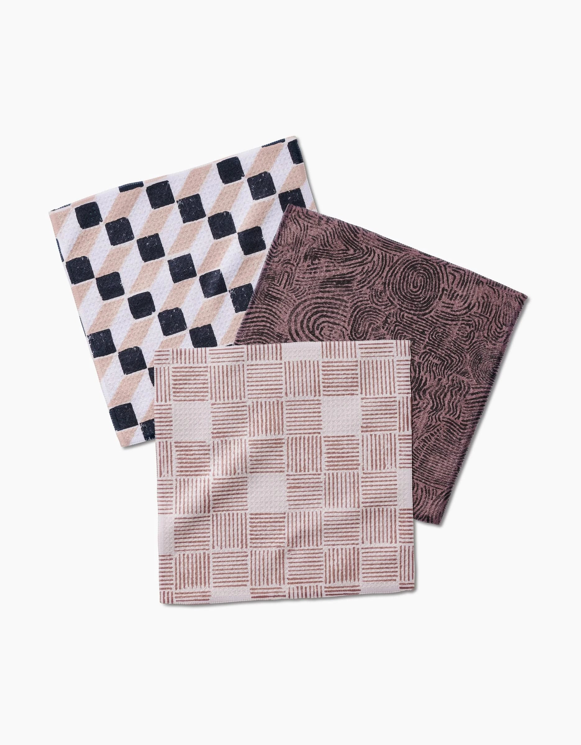 Three Chiseled cloth napkins by Studio Jeannot, made from recycled materials, are arranged overlapping: one has a black and beige 3D cube design, another a dark brown swirl motif, and the third shows a beige and white checkered pattern.