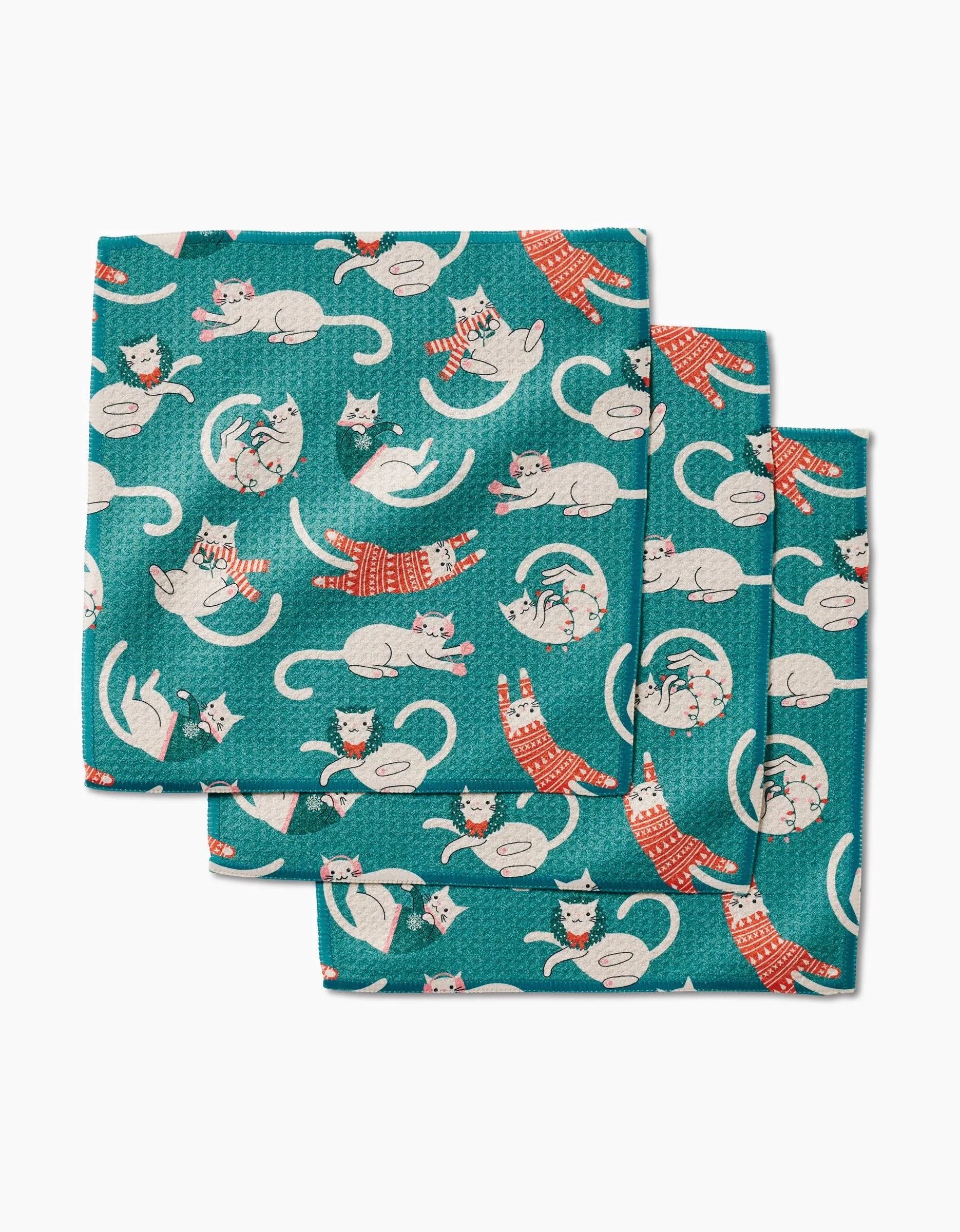 The Christmas Cats napkin set by Megan Kampa includes three teal cloth napkins made from recycled materials, with a playful white cat pattern—some wearing red sweaters, others napping or stretching in fun poses.