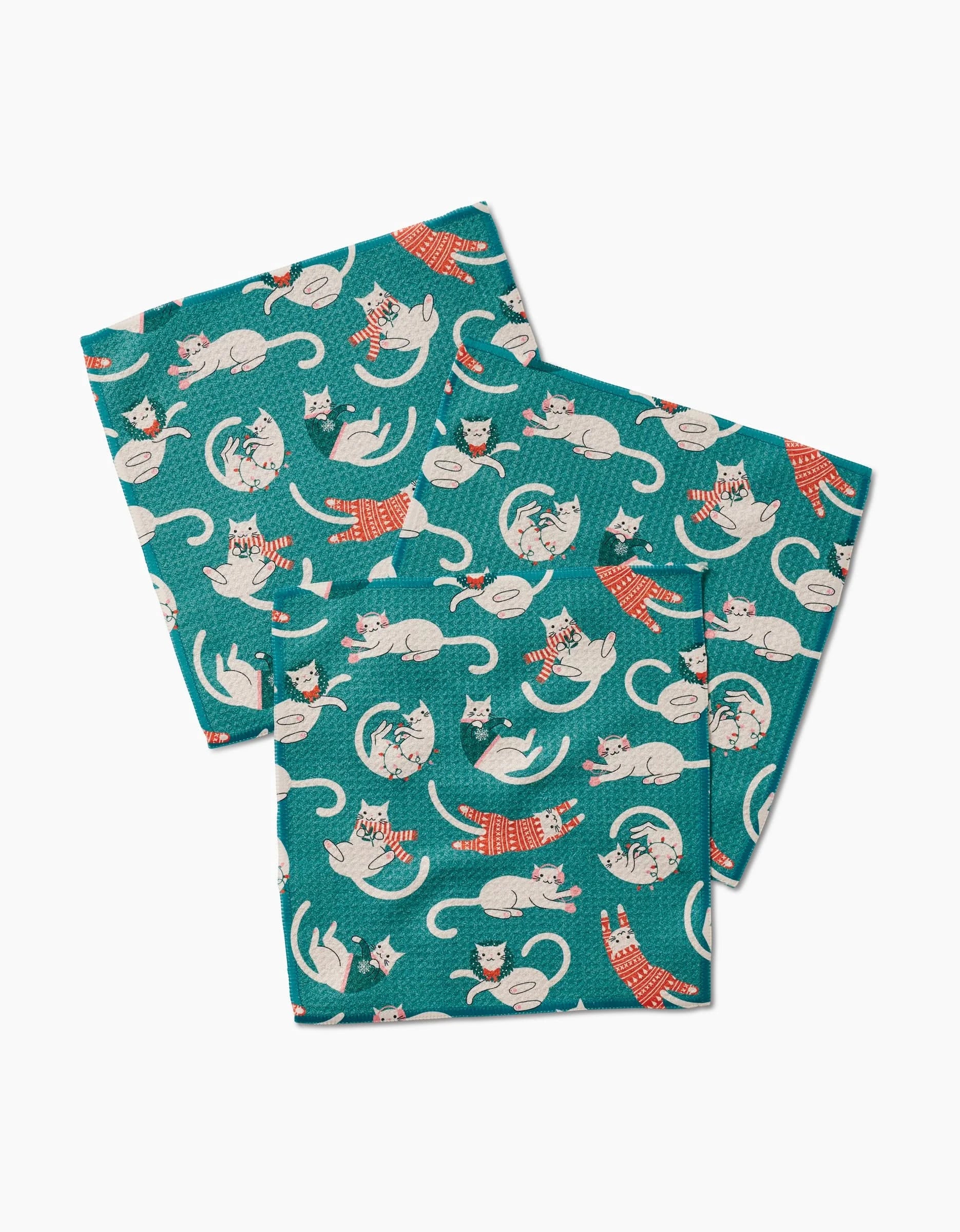 The Christmas Cats dishcloths by Megan Kampa feature a playful pattern of white and orange cats in red striped scarves, set on teal recycled fabric. The illustrated cats appear in various poses across the overlapping cloths.