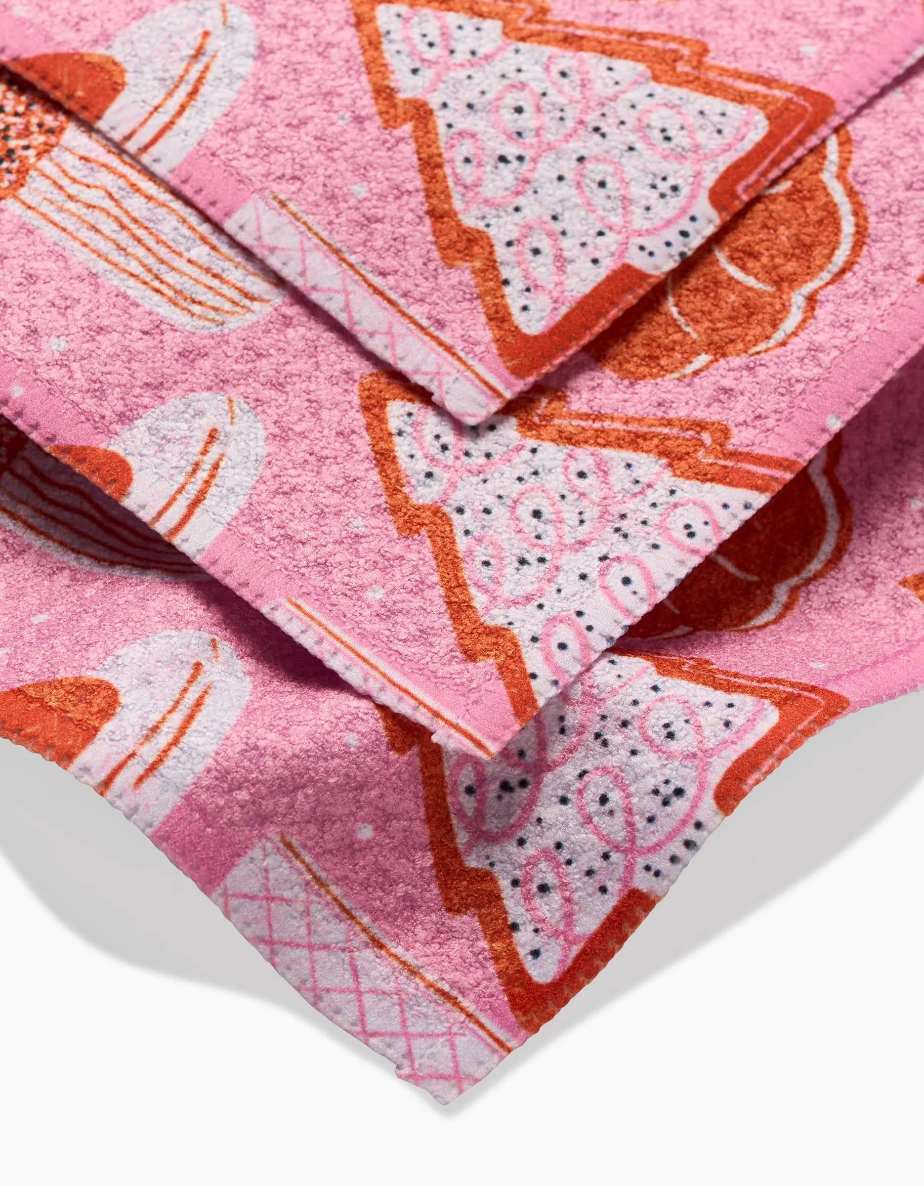 Close-up of folded pink Megan Roy Christmas Cookies Geoweave dishcloths, featuring whimsical cake and pie slice illustrations in white, red, and orange with decorative patterns.
