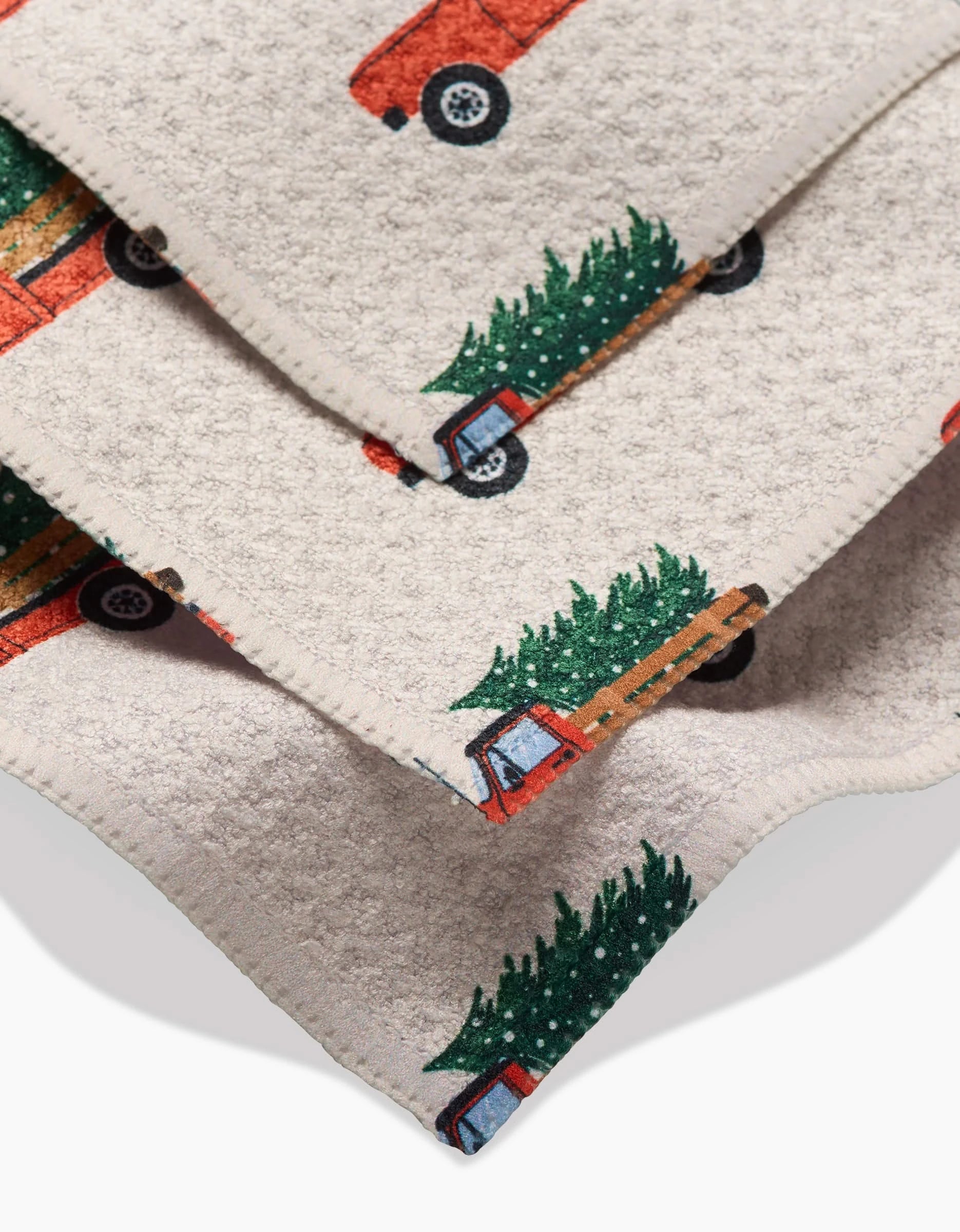 Close-up of the Christmas Ride dishcloth by Megan Kampa, made of beige Geoweave fabric with red trucks carrying green Christmas trees printed on it, neatly stacked with stitched edges.