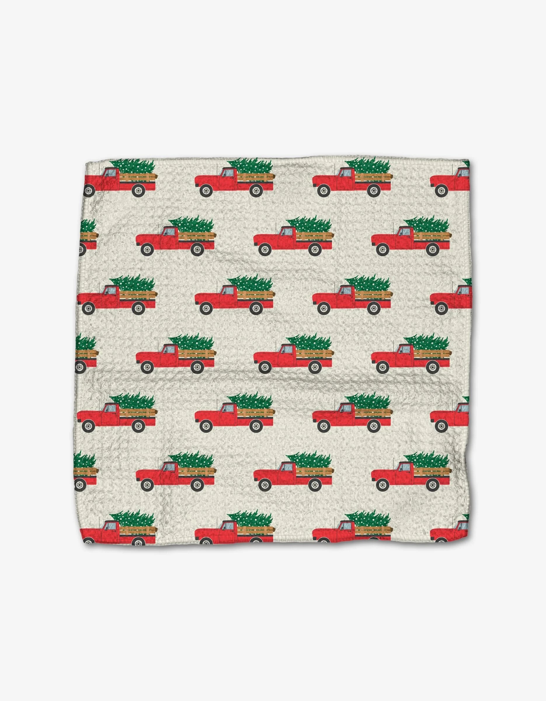 The Christmas Ride cloth napkin by Megan Kampa features Geoweave fabric with a repeating pattern of red pickup trucks carrying green Christmas trees on a light background.