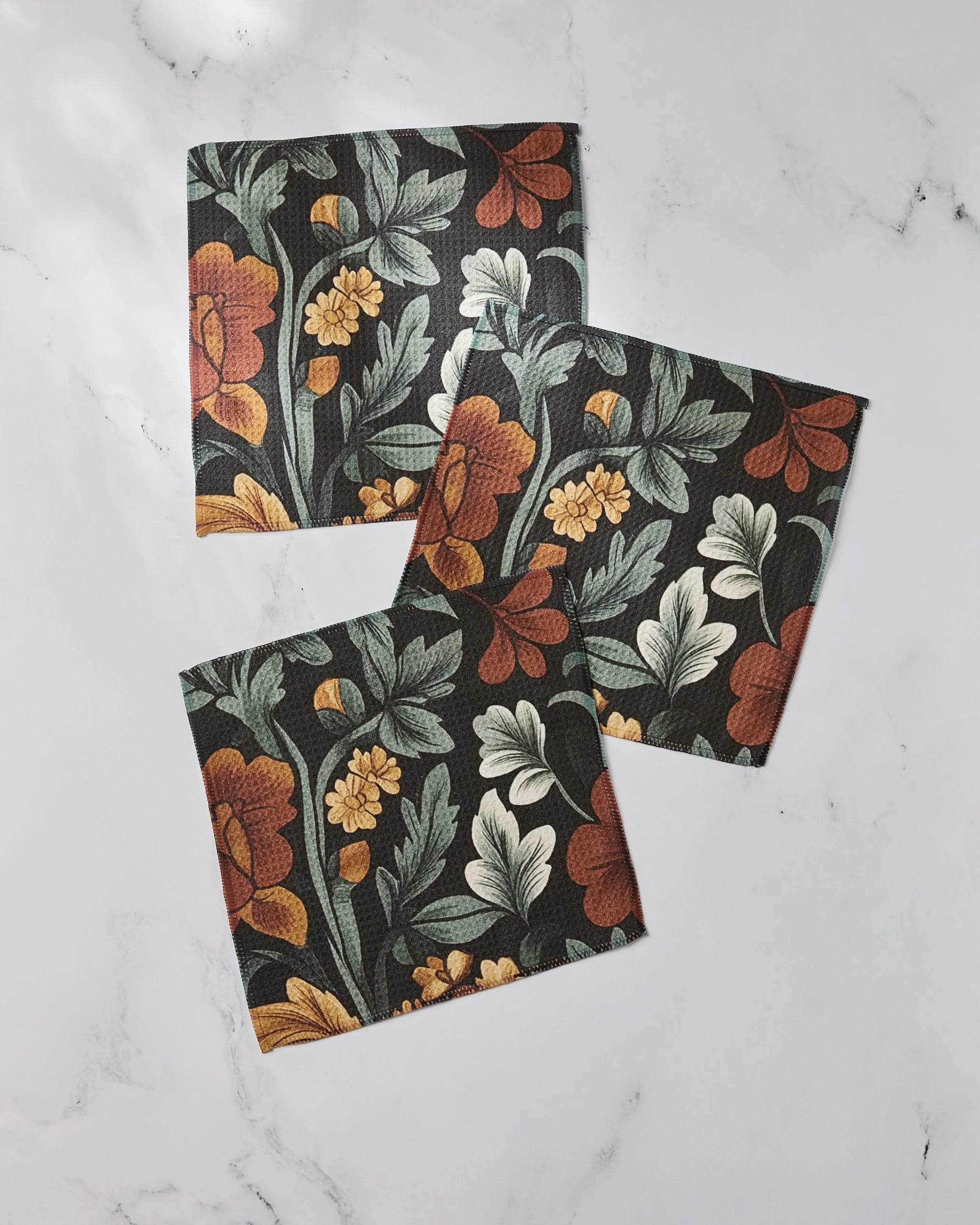 Three Classic Floral napkins from Geometry, made with recycled materials, showcase orange, yellow, and cream flowers with green leaves on a dark background, elegantly displayed on white marble.
