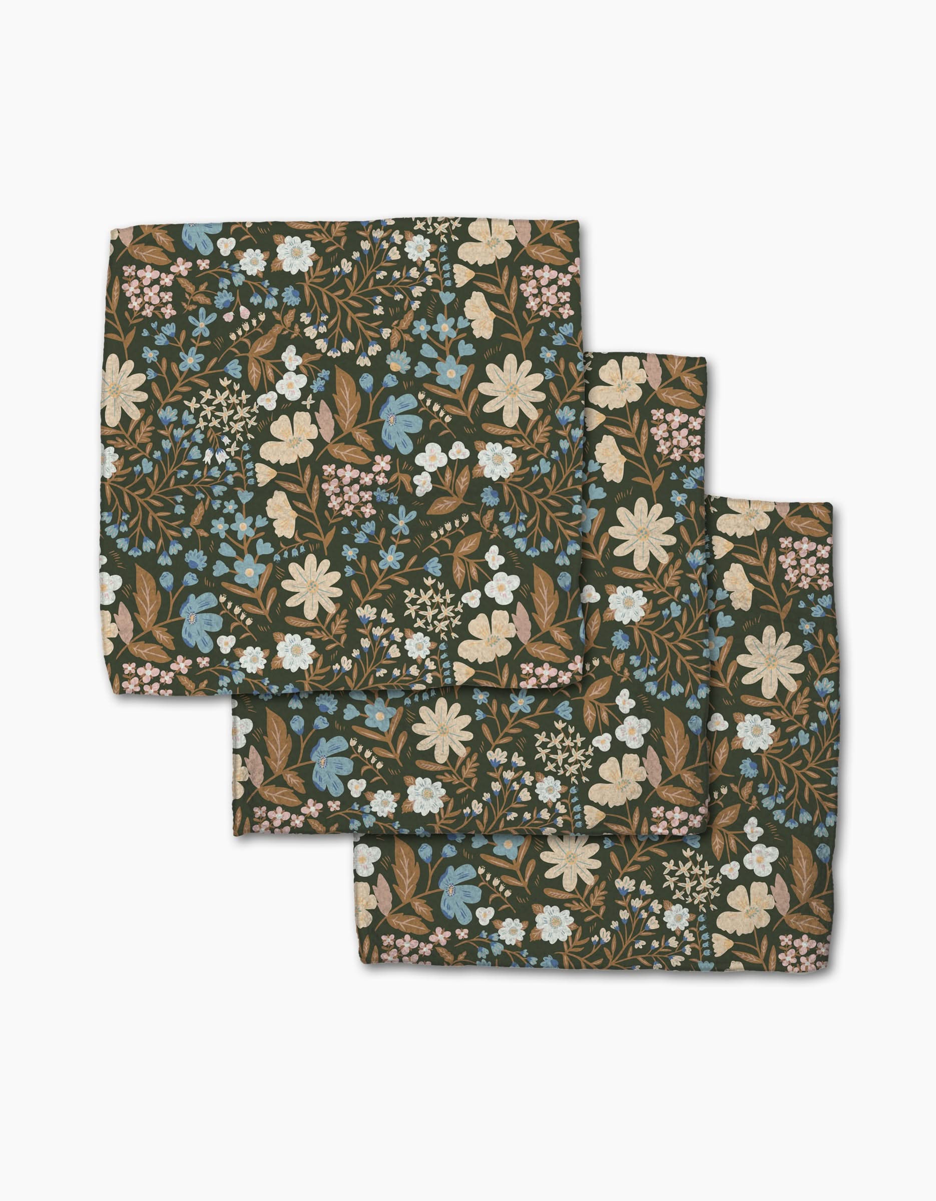 Three folded Cottage Meadow Sepia cloth napkins from Danelys Sidron, made of Geoweave fabric with a floral print in white, blue, and beige flowers plus green leaves on a dark background, are arranged in a layered stack.