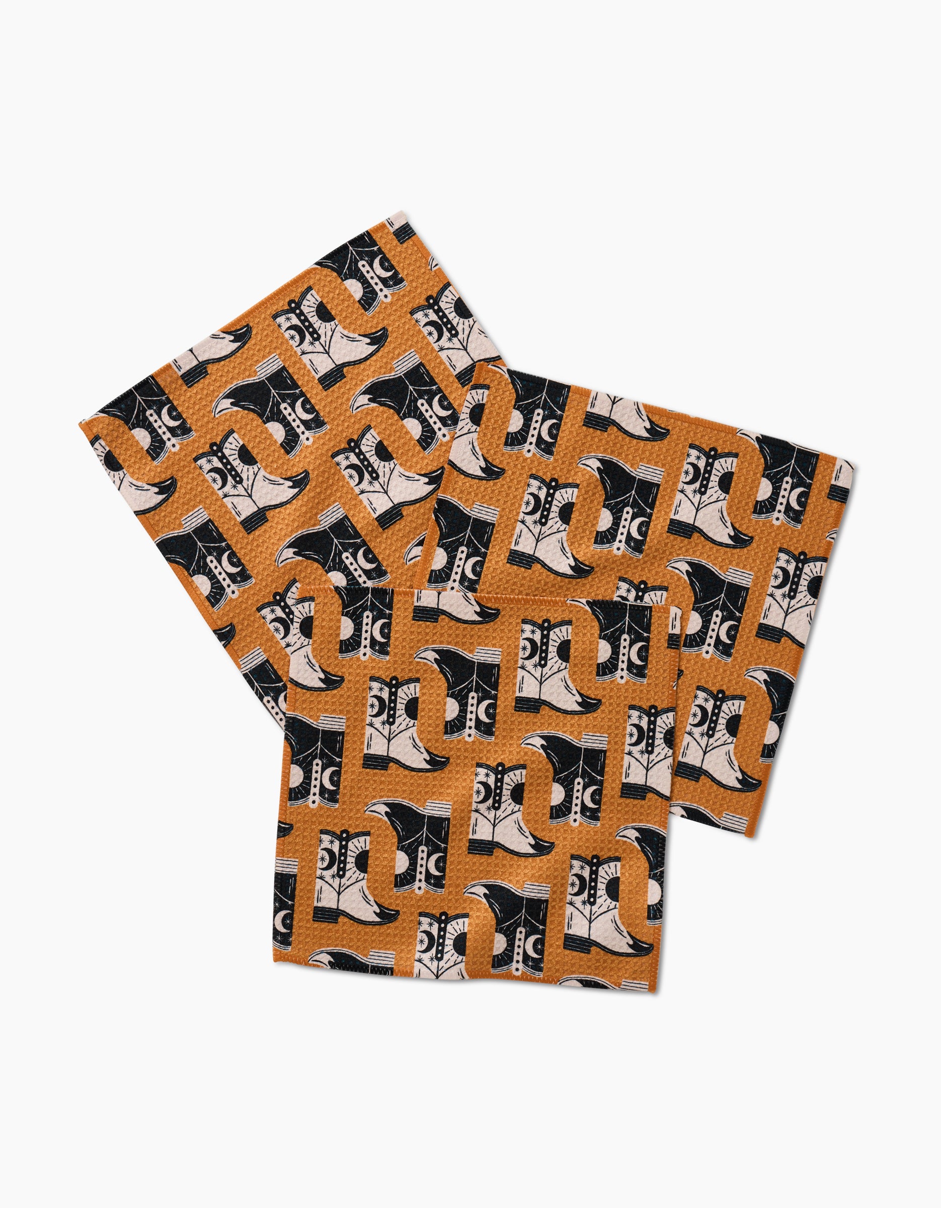 Two rectangular Megan Roy Geoweave dishcloths in the Cowboy Boots pattern, featuring orange backgrounds with black and white cowboy boots, are slightly overlapping on a white surface.