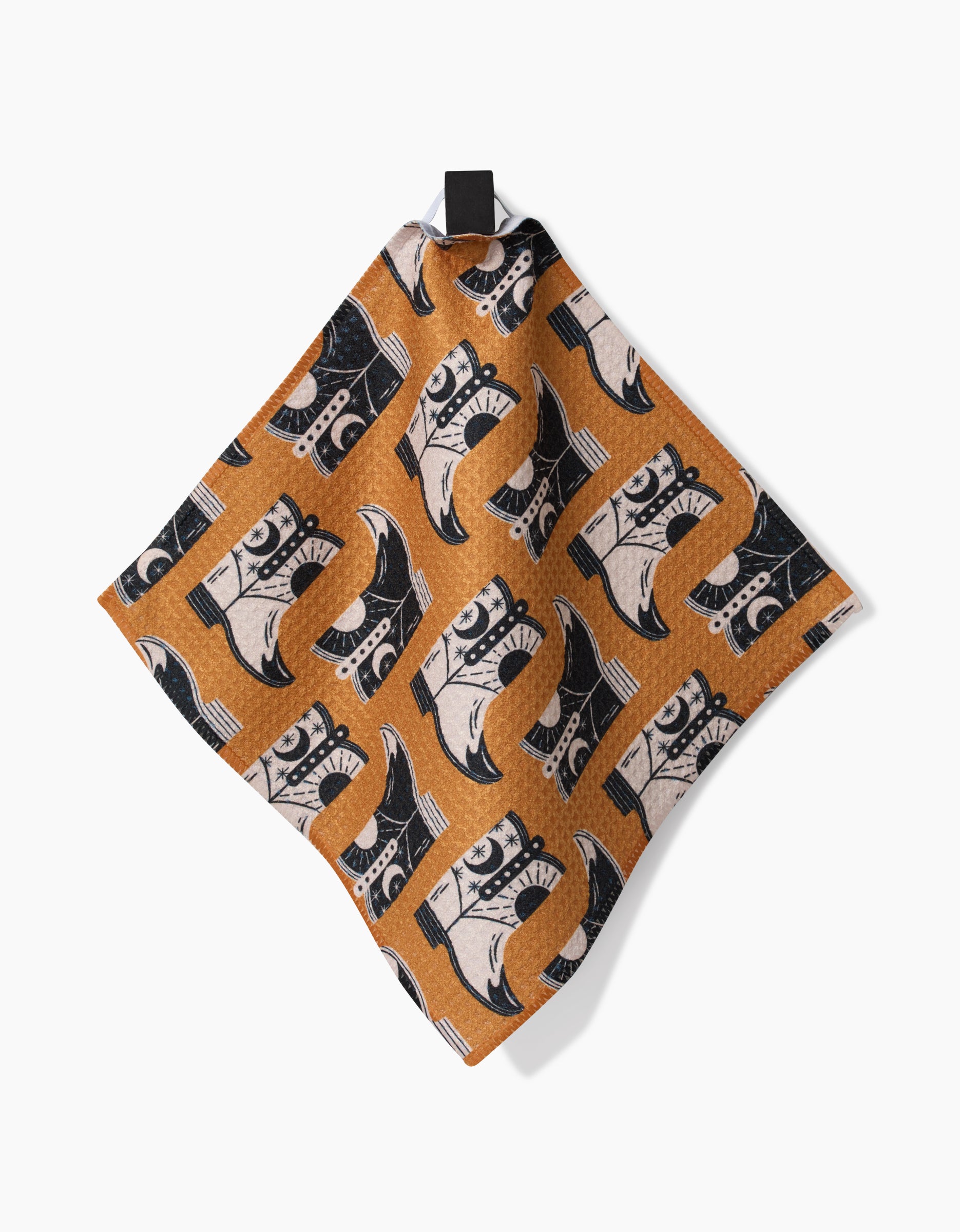 The Megan Roy Cowboy Boots kitchen towel, made from recycled materials, displays a brown base with a black and white cowboy boot pattern and hangs by a black loop against a plain white background.