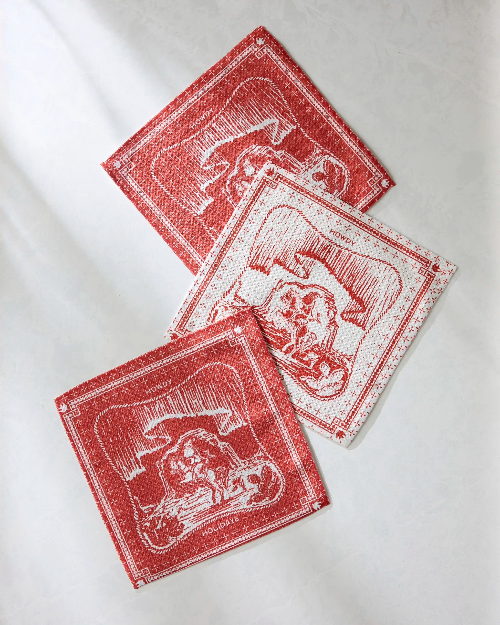 Three Geometry Cowboy Christmas napkins, made from recycled materials and featuring red and white lion and crown motifs, are arranged at varying angles on a light textured surface with some overlap.