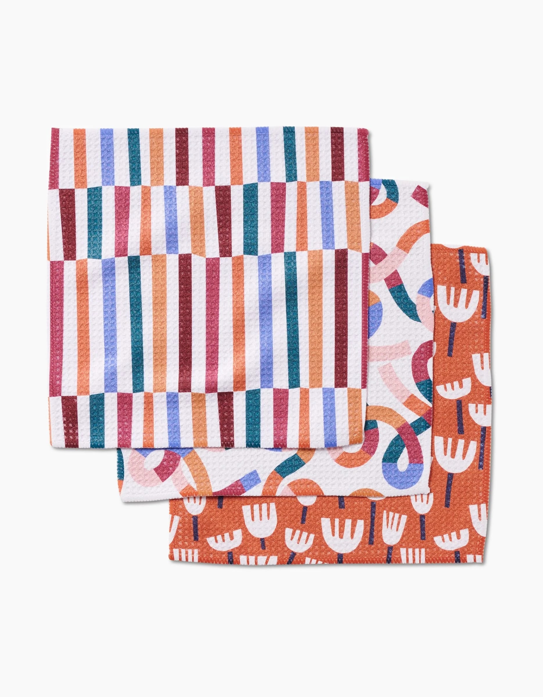 Three folded dish towels from Claire Iglesiass Cutout Shapes set: one with vibrant stripes, one with abstract loops, and one with red and white tulip-like shapes—all made from premium Geoweave fabric for style and absorbency.