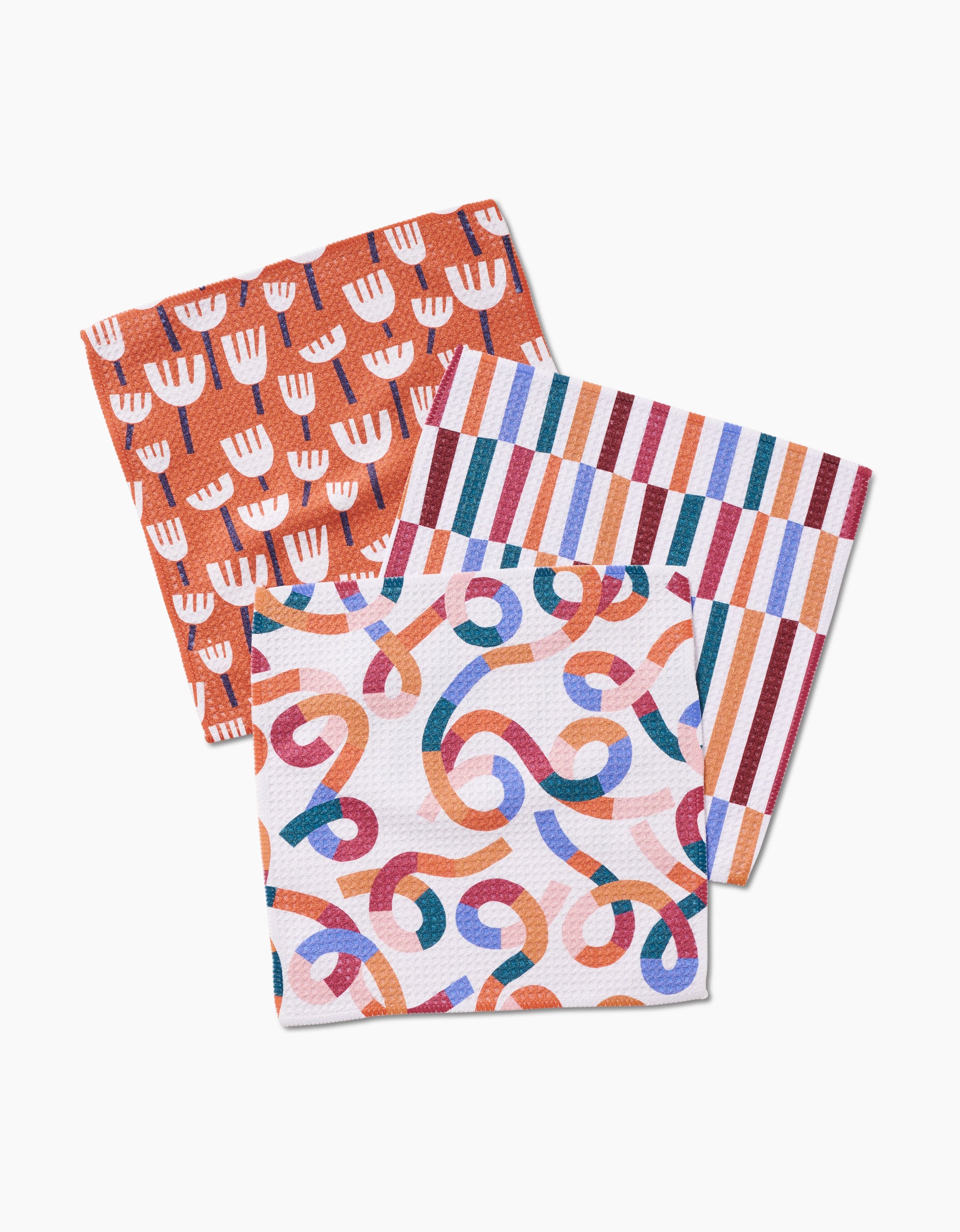 The Claire Iglesias Cutout Shapes Dishcloth Set includes three rectangular cloths with vibrant patterns—white flowers on orange, multicolored stripes, and swirling designs—crafted with Geoweave fabric for stylish, super-absorbent kitchen use.