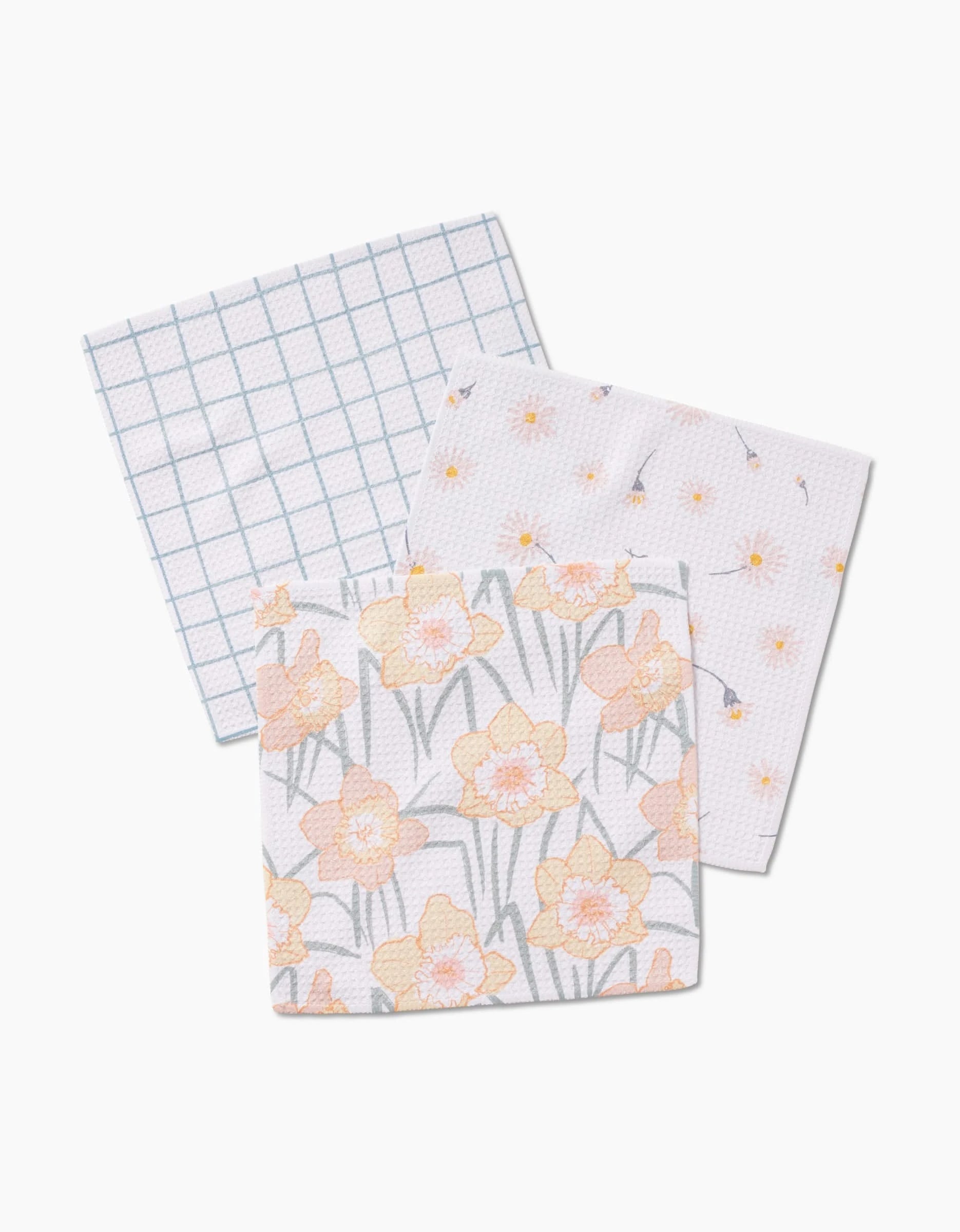 The Hufton Studio Daisy Dust set includes three square dishcloths: one with a blue grid, one with small yellow flowers, and one with large yellow daffodils on white, all crafted from super-absorbent Geoweave fabric.