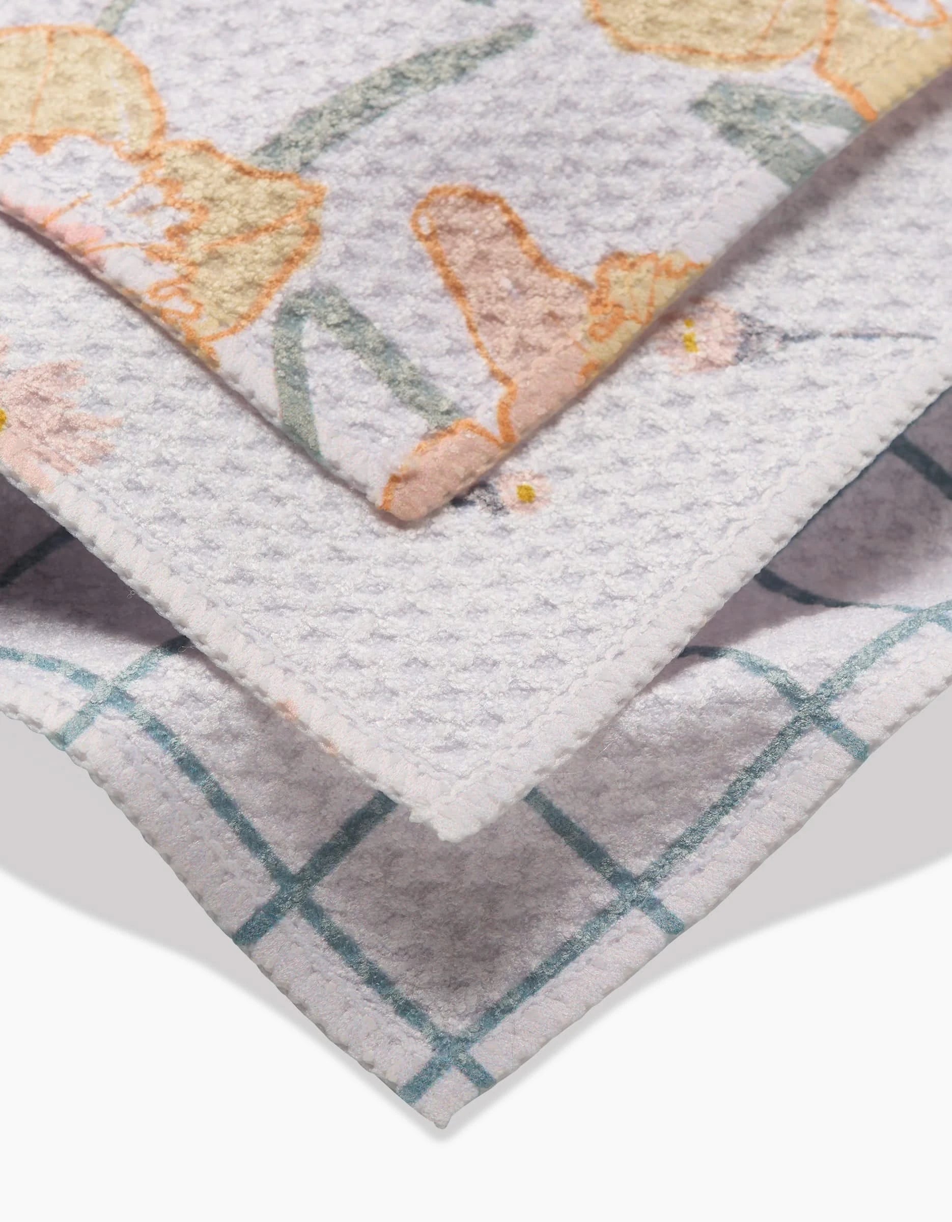 Close-up of three stacked Hufton Studio Daisy Dust dishcloths: a floral pattern in soft orange and green, a plain light grey, and a teal grid on white Geoweave fabric for super-absorbent cleaning.