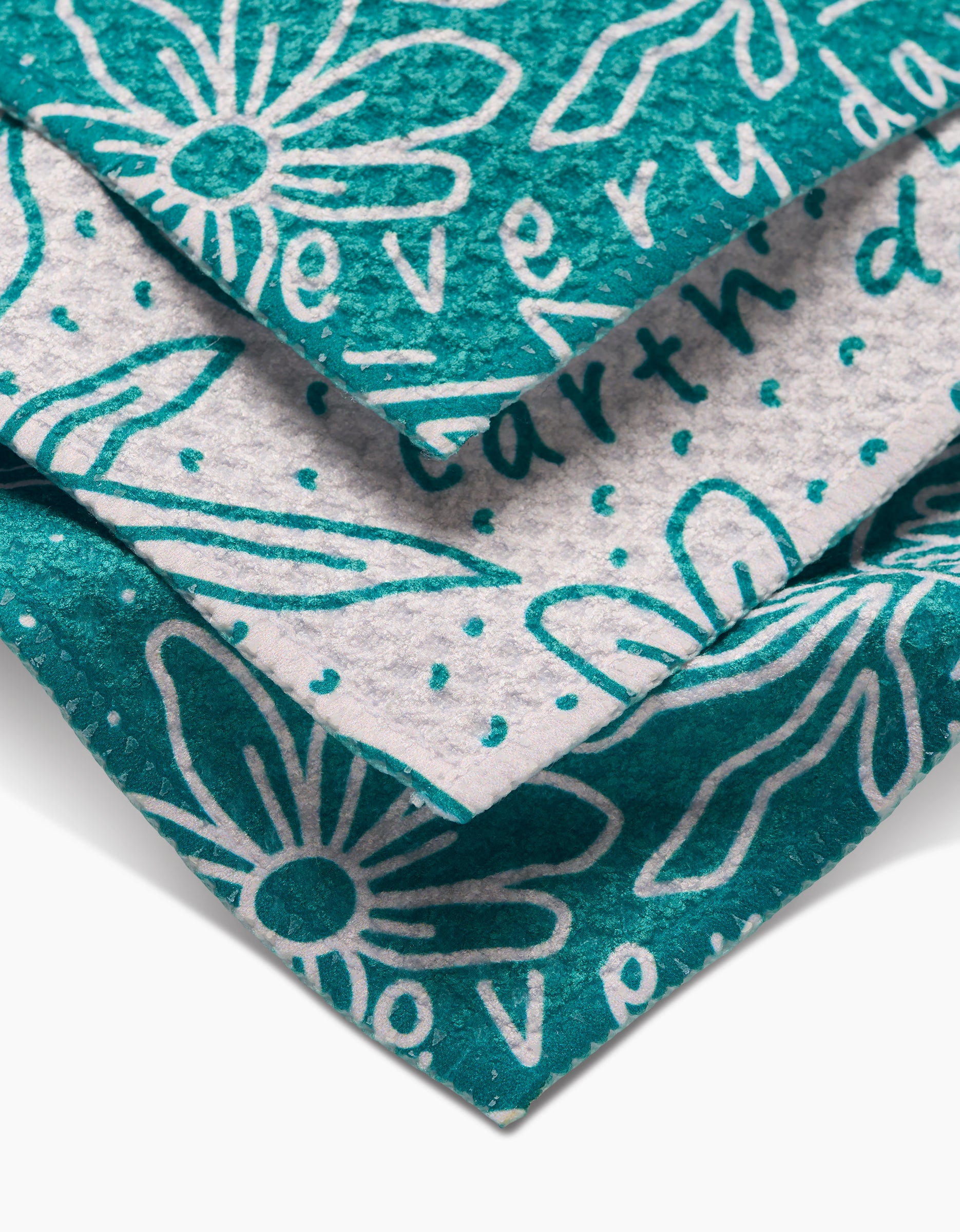 Close-up of stacked GEOMETRY Every Day Geoweave fabric dishcloths in teal and white, with hand-drawn daisy designs and partially visible handwritten phrases. Fabric texture is clearly visible.