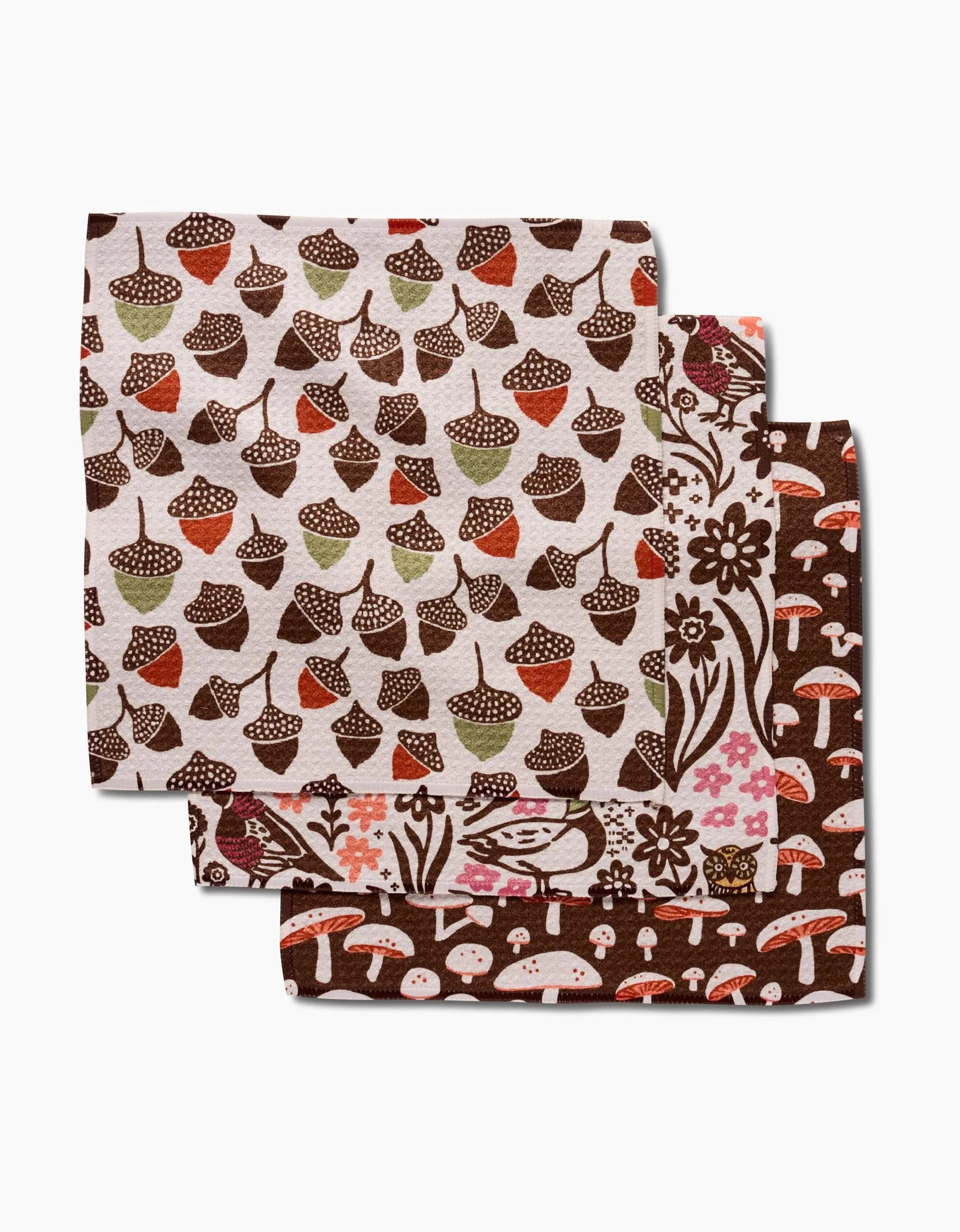 The Fall Abundance napkins by Kayla Ann include three folded cloths made from recycled materials, featuring autumn patterns: earthy acorns, brown florals with mushrooms, and assorted mushrooms on a dark brown background.