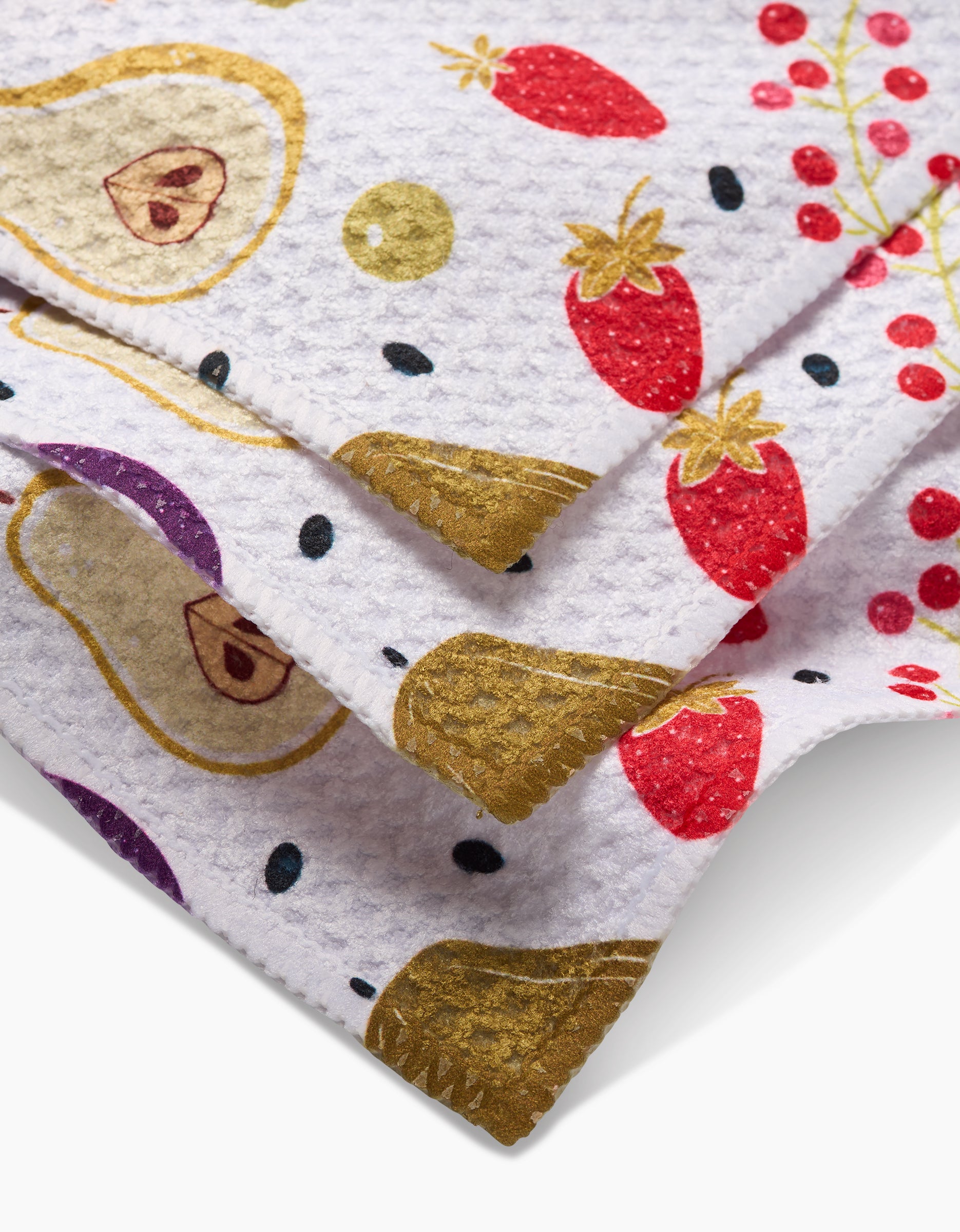 A close-up of the Sara Mari Farmers Market dishcloth shows its textured white Geoweave fabric with colorful prints—strawberries, red berries, purple and yellow fruits, and gold circles—neatly layered to highlight its quality weave.