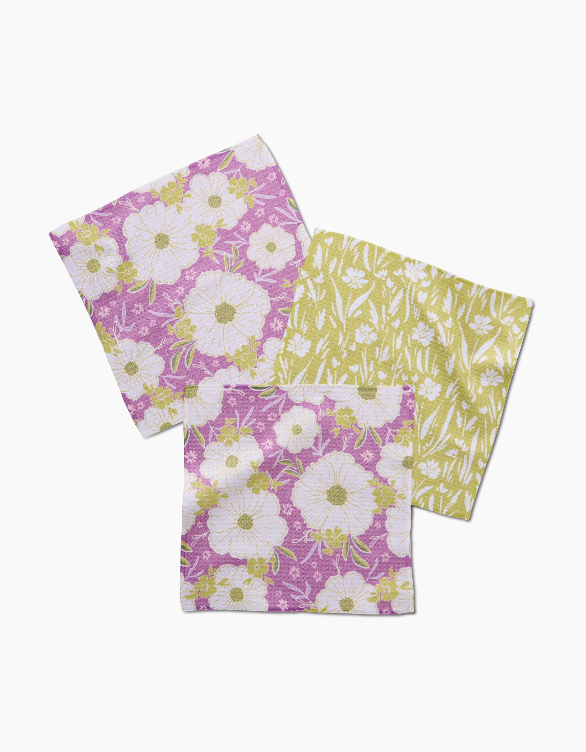 Three “Field of Spring Flowers” square cloth napkins by Ashley Celena—two pink and white with large flowers, one yellow and white with small florals—all made from recycled materials, are arranged overlapping on a white background.