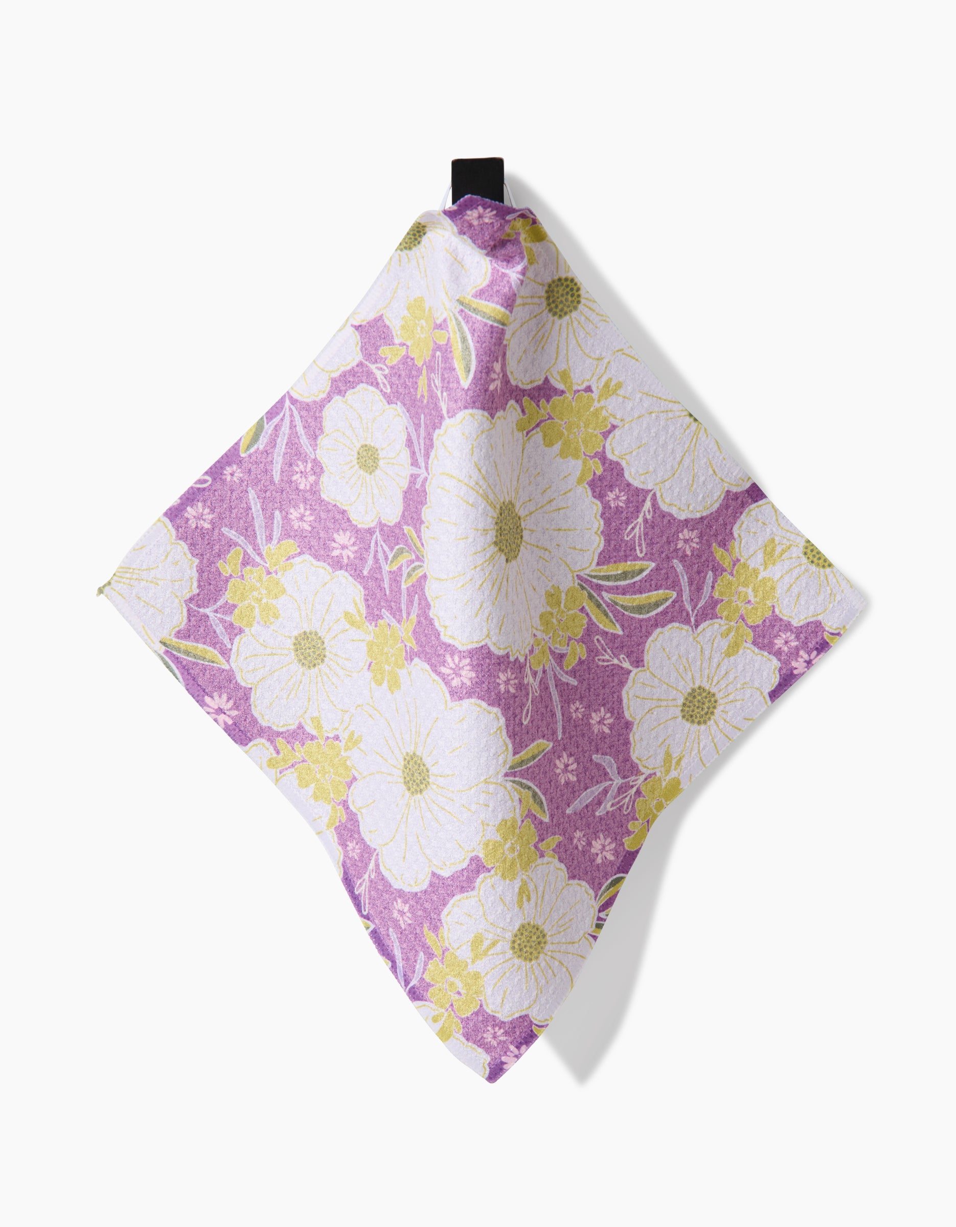 The Field of Spring Flowers napkin by Ashley Celena is made from Geoweave fabric, featuring a purple background with large white and small yellow flowers, and hangs from a black loop at the top.