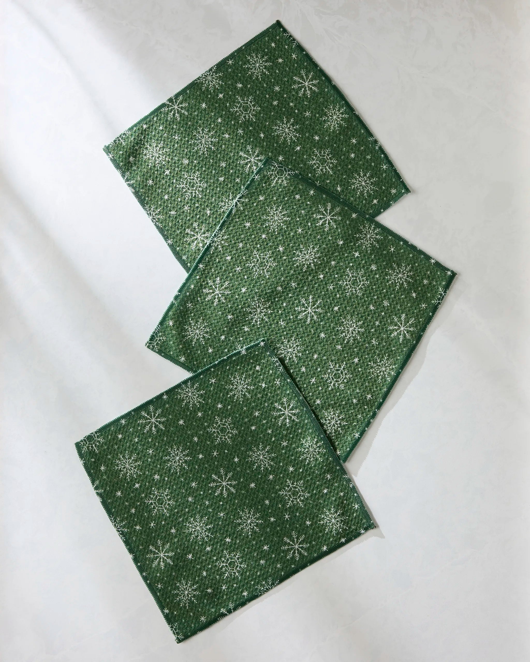 Three Geometry Firlight green square napkins, featuring white snowflake patterns and made from recycled materials, are arranged diagonally on a light surface.