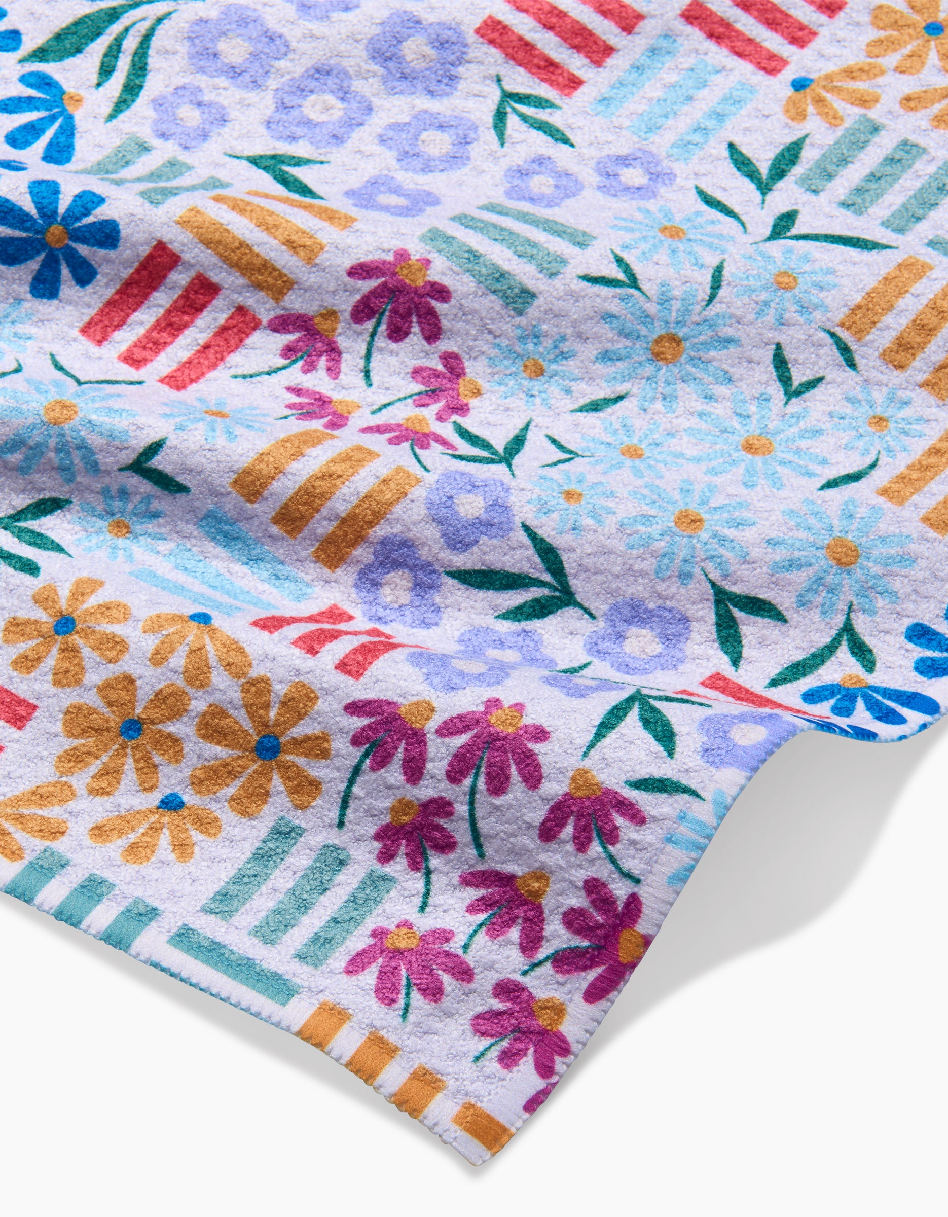 A close-up of Charlie Rowan Designs Floral Garden dishcloth shows colorful flowers and stripes in blue, purple, orange, and pink on white. Made from recycled materials, the towel is slightly folded at one corner.