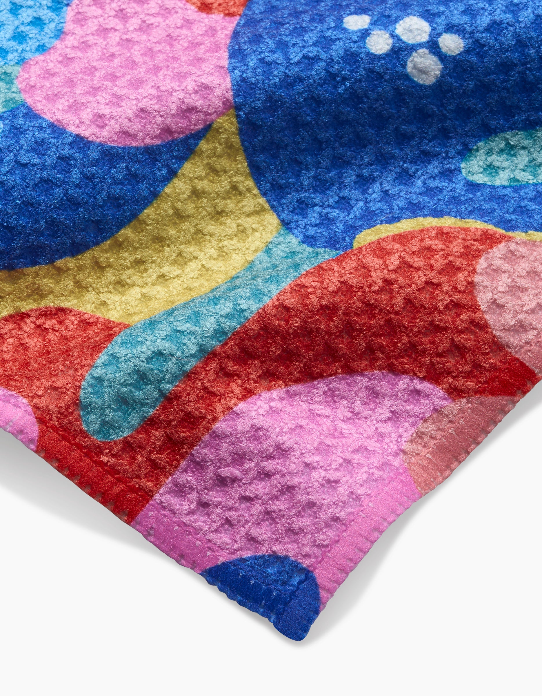 A close-up of the Masha Volnova Flower Powers dishcloth shows its textured Geoweave fabric with a colorful abstract pattern of blue, red, pink, yellow, and teal organic shapes plus white circles.