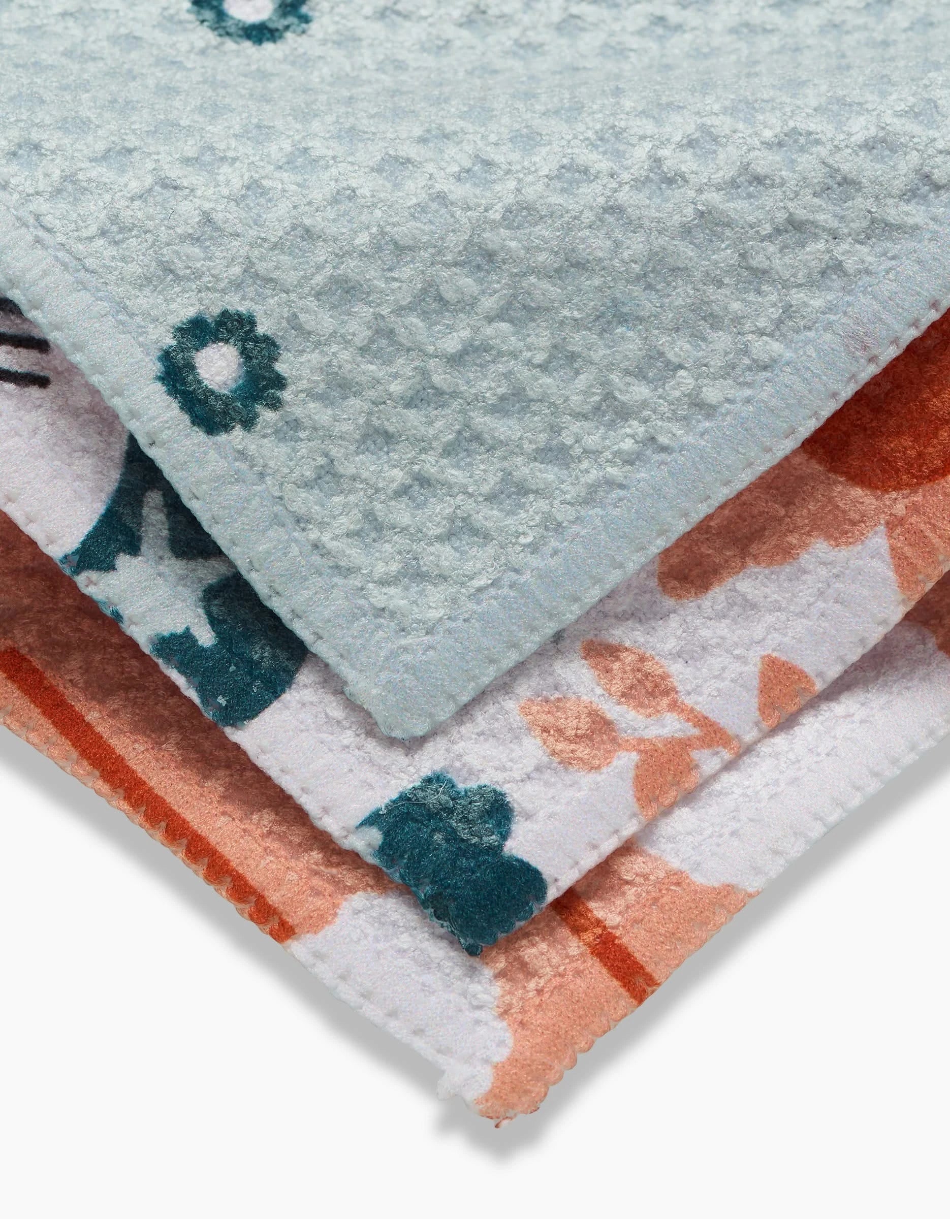 Three folded Coit Creative Forest Florals towels are stacked, each with unique textures and patterns. The top is light blue with a honeycomb texture and dark florals; the rest have blue, orange, and white floral designs using recycled materials.