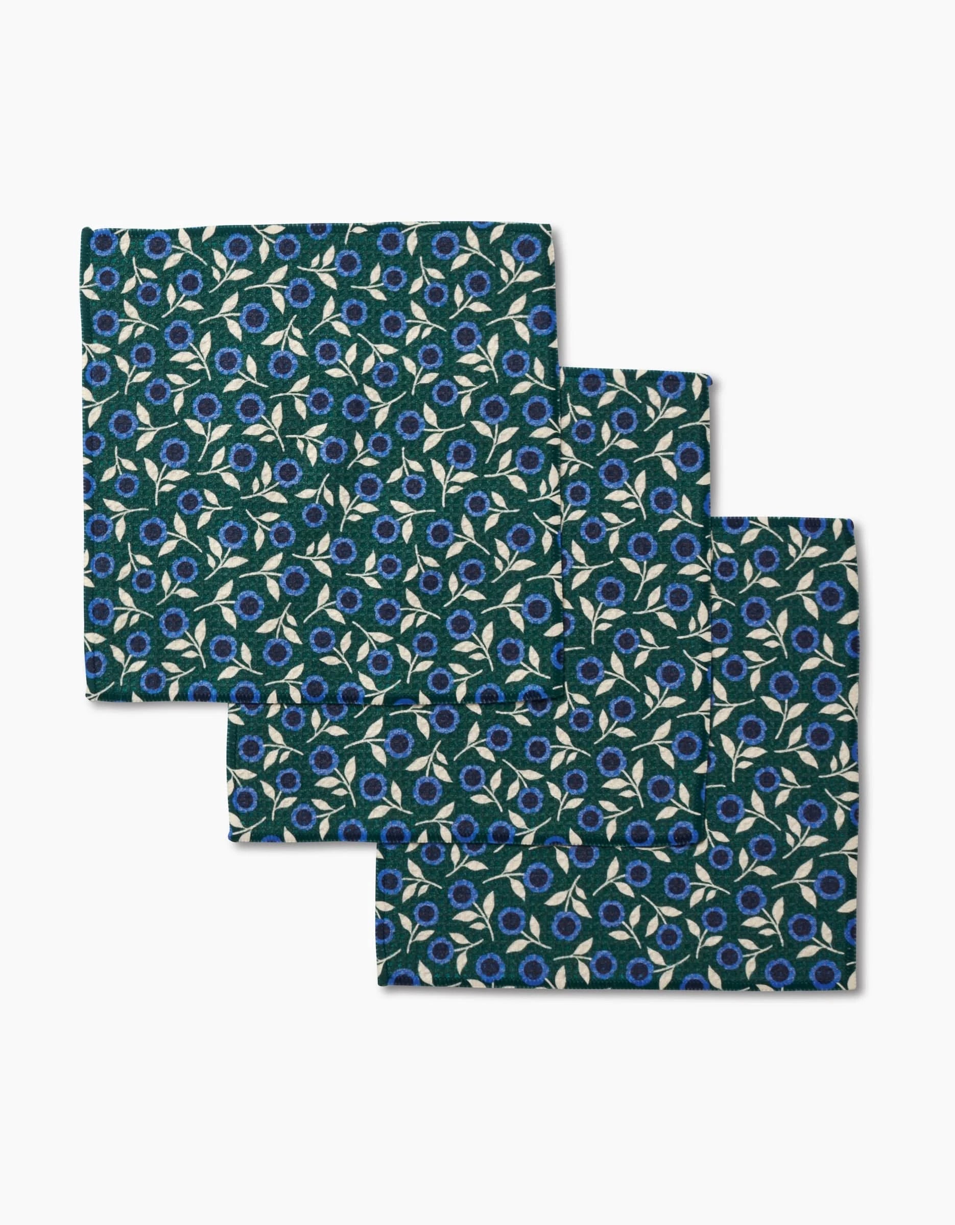 Three neatly folded Megan Kampa Friendly Fleurs pocket squares with a dark green floral print rest on a plain white surface, crafted from Geoweave fabric.