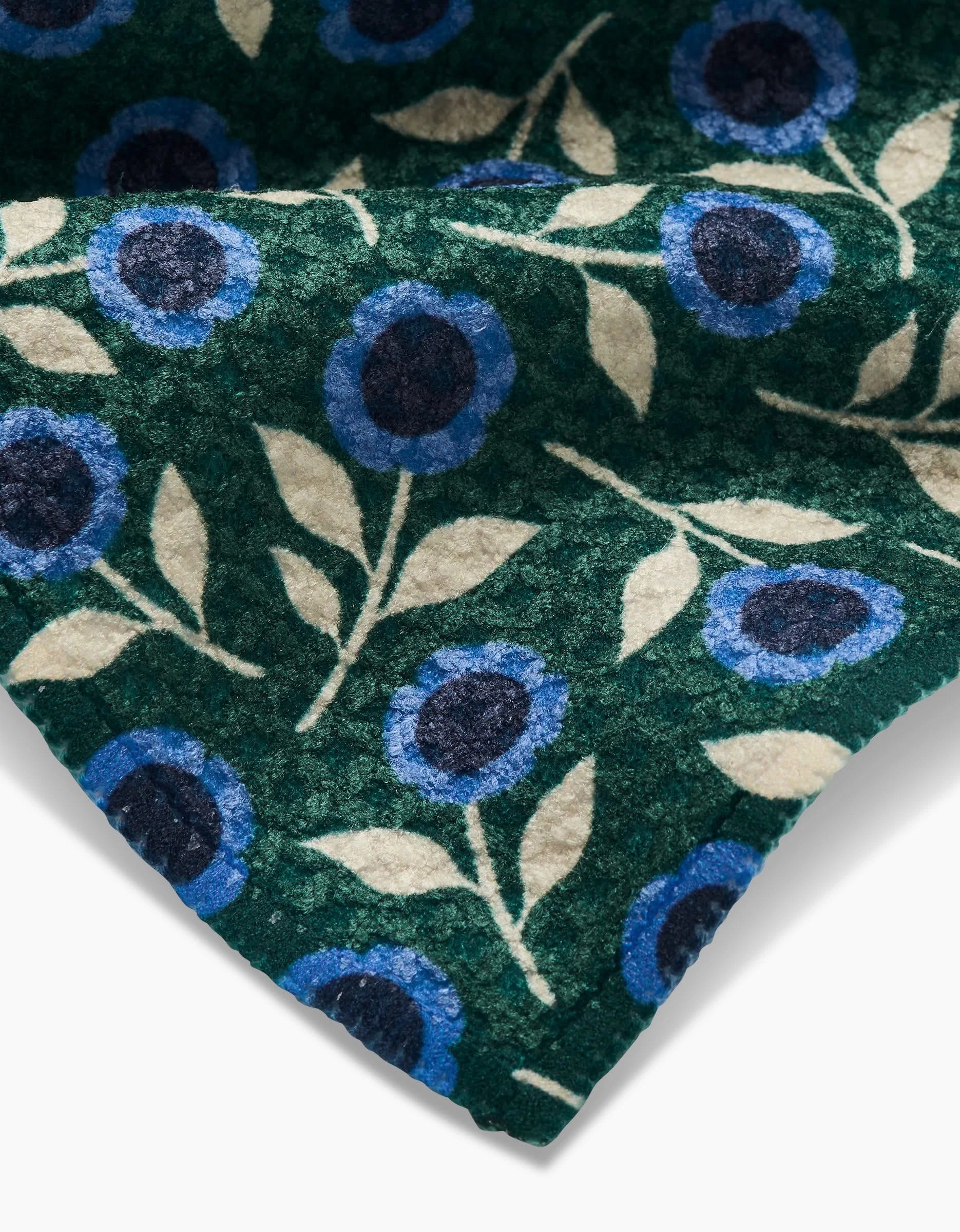A close-up of Friendly Fleurs by Megan Kampa shows Geoweave fabric with blue flowers, cream leaves, and a green background. The softly folded texture highlights its detail—ideal for chic dishcloths.