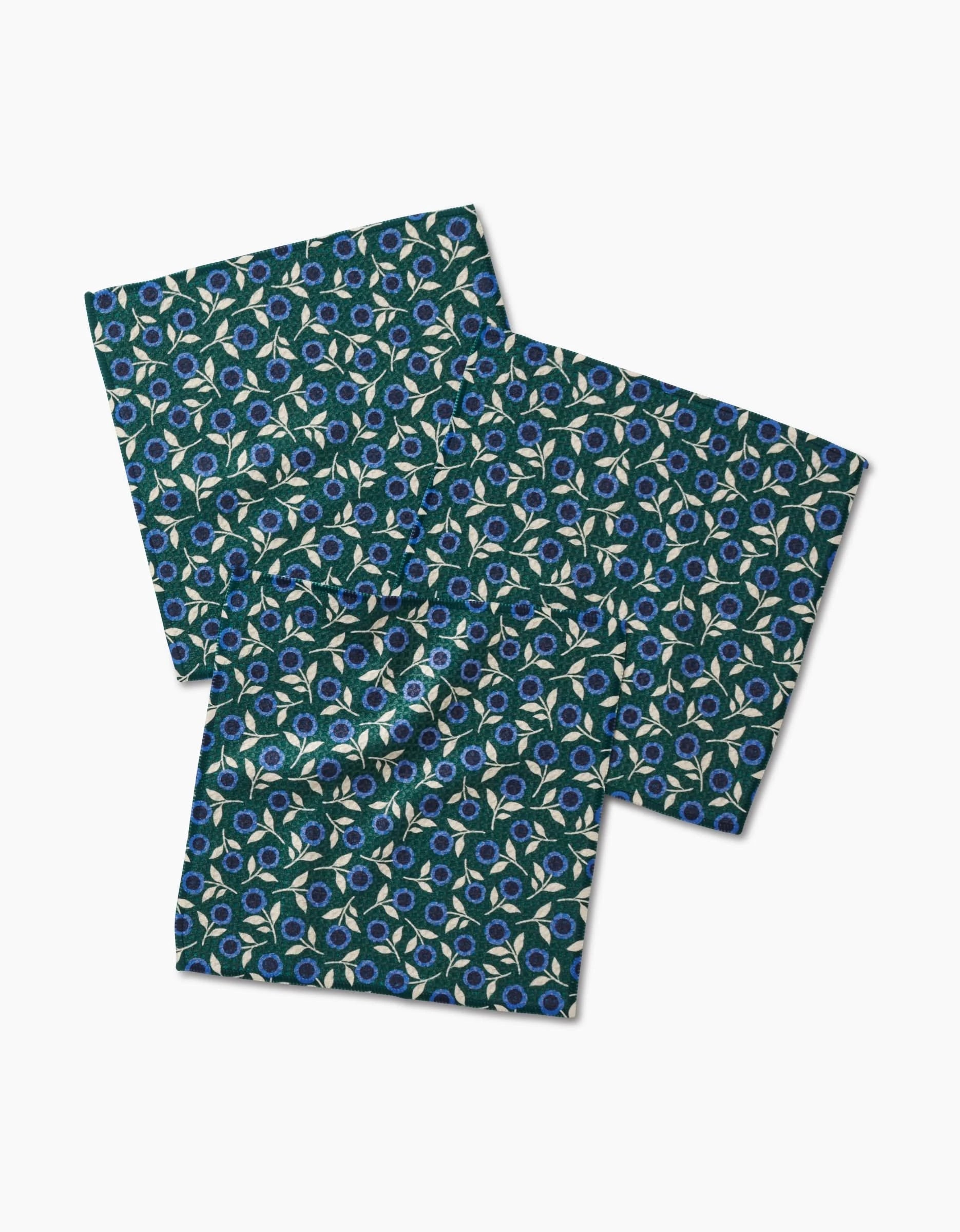 Three Friendly Fleurs square dishcloths by Megan Kampa, made from recycled materials, show a repeating blue, green, and white floral pattern on a dark background. They are arranged overlapping on a white surface.