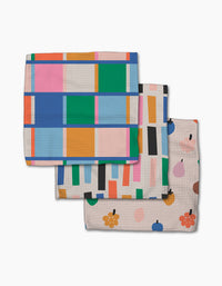 Three Jennifer Bouron Fruit Basket Geoweave dishcloths stacked in a fan: the top has a colorful checkered grid, the middle shows multicolored vertical rectangles, and the bottom features abstract fruit and flower illustrations.